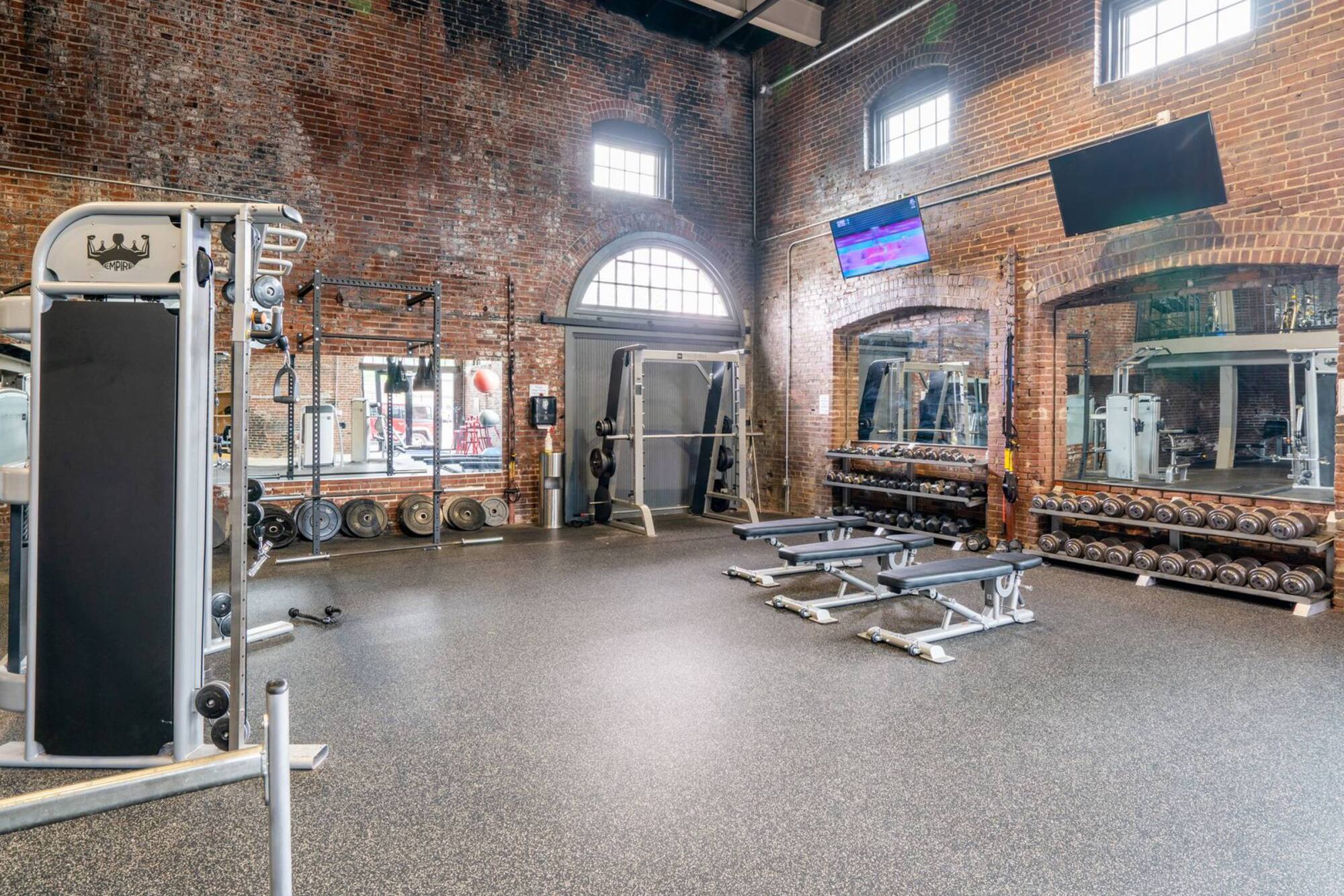 Treadmills and elliptical machines in a gym with brick walls and large windows, viewed from an upper level.