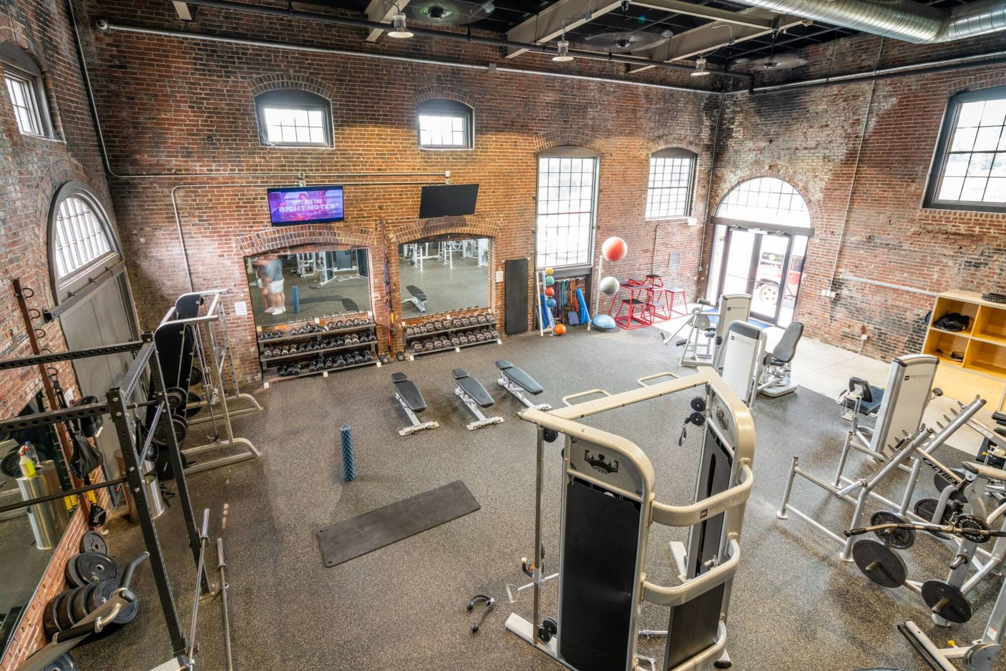 Spacious gym with weight machines, benches, free weights, exercise balls, and large windows in a brick building.