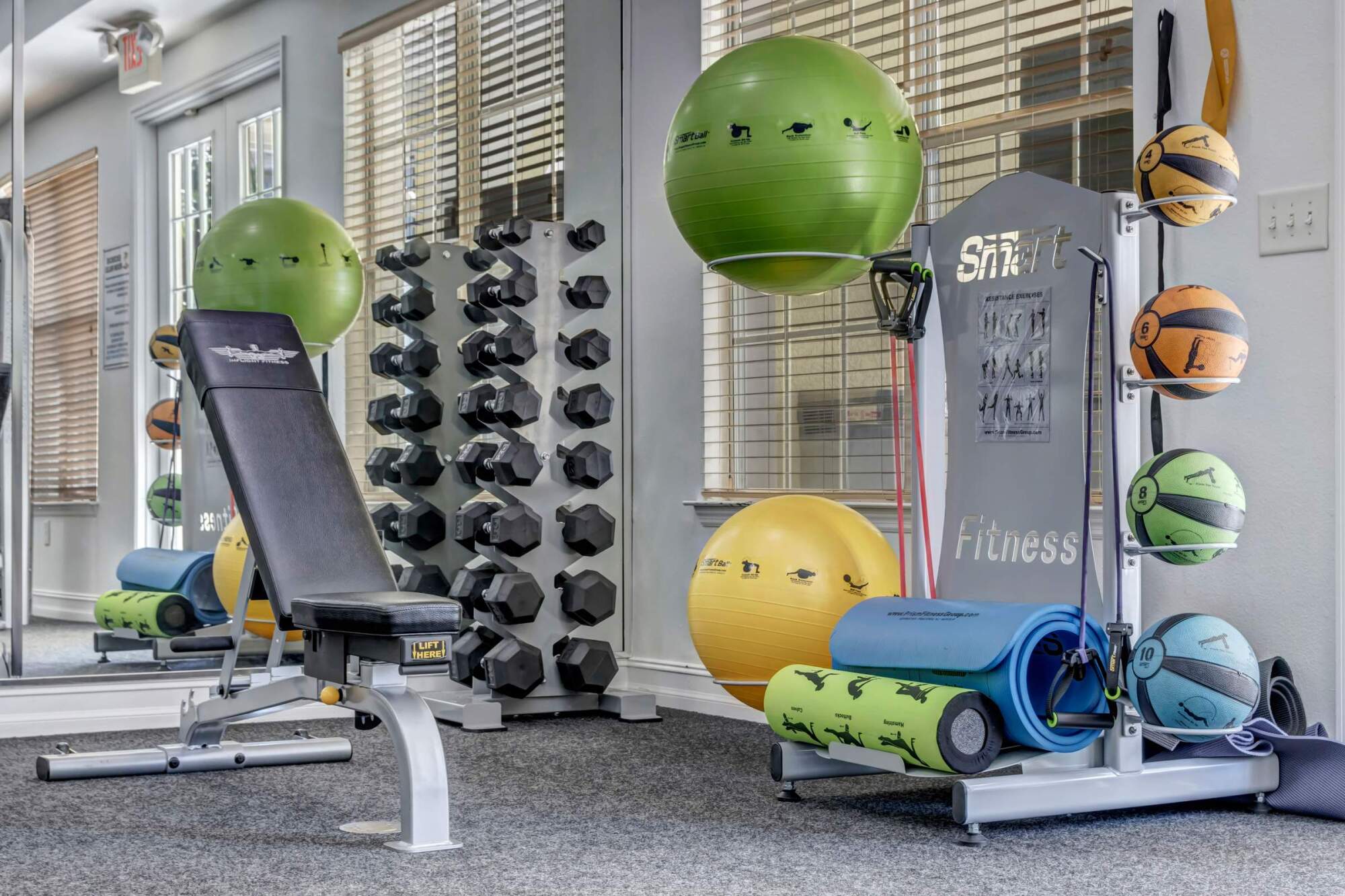 Apartment gym with a weight bench, dumbbells, exercise balls, yoga mats, and medicine balls near mirrored walls.