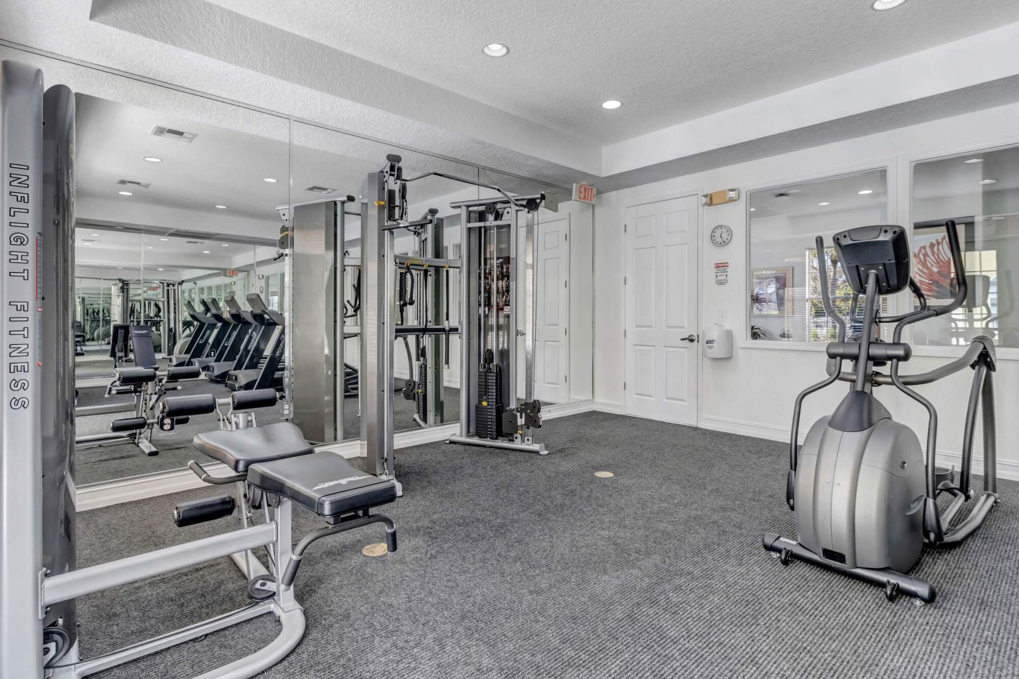 Small gym room with strength and cardio equipment, mirrored wall, gray carpet, and white walls.