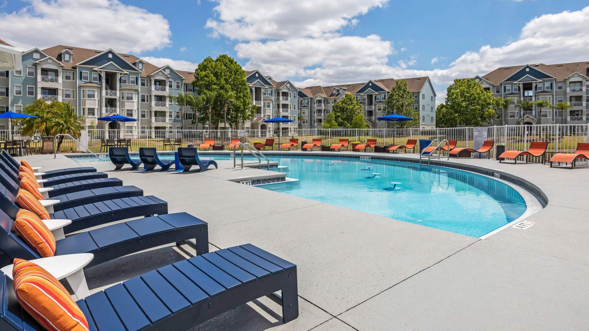 Outdoor pool with blue lounge chairs and orange pillows, surrounded by apartment buildings under a partly cloudy sky.