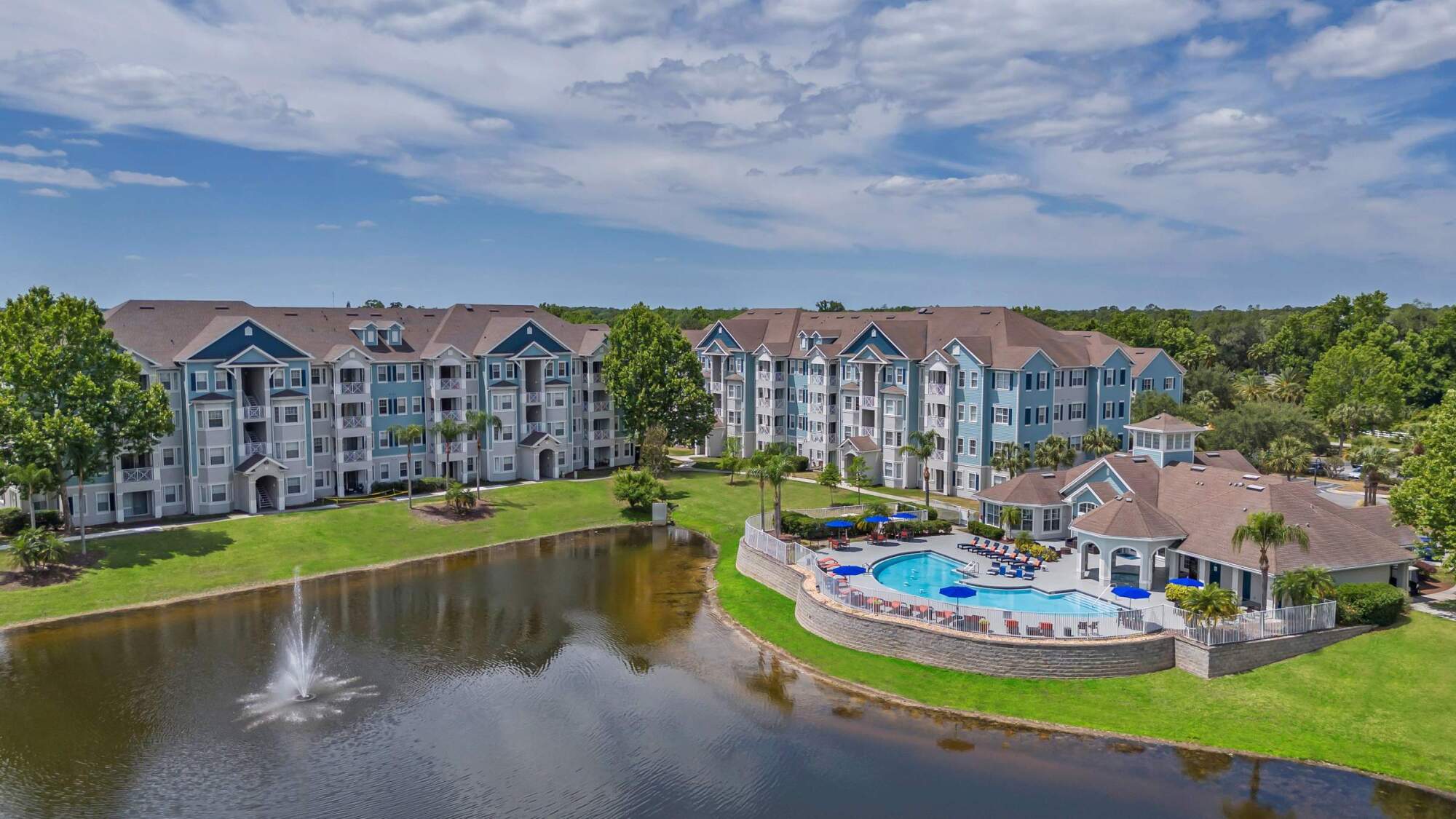 Aerial view of apartment buildings near a pond with a fountain and a swimming pool surrounded by lounge chairs.
