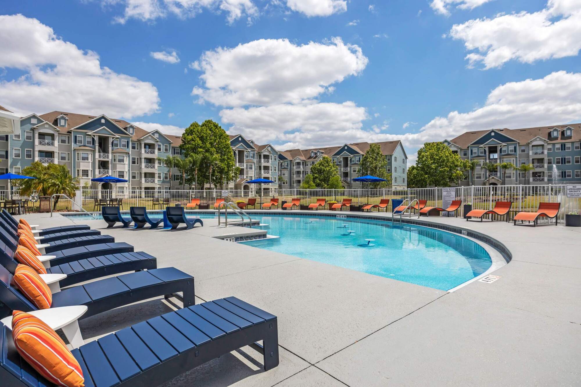 Outdoor apartment pool area with lounge chairs, umbrellas, and modern buildings in the background under a blue sky.