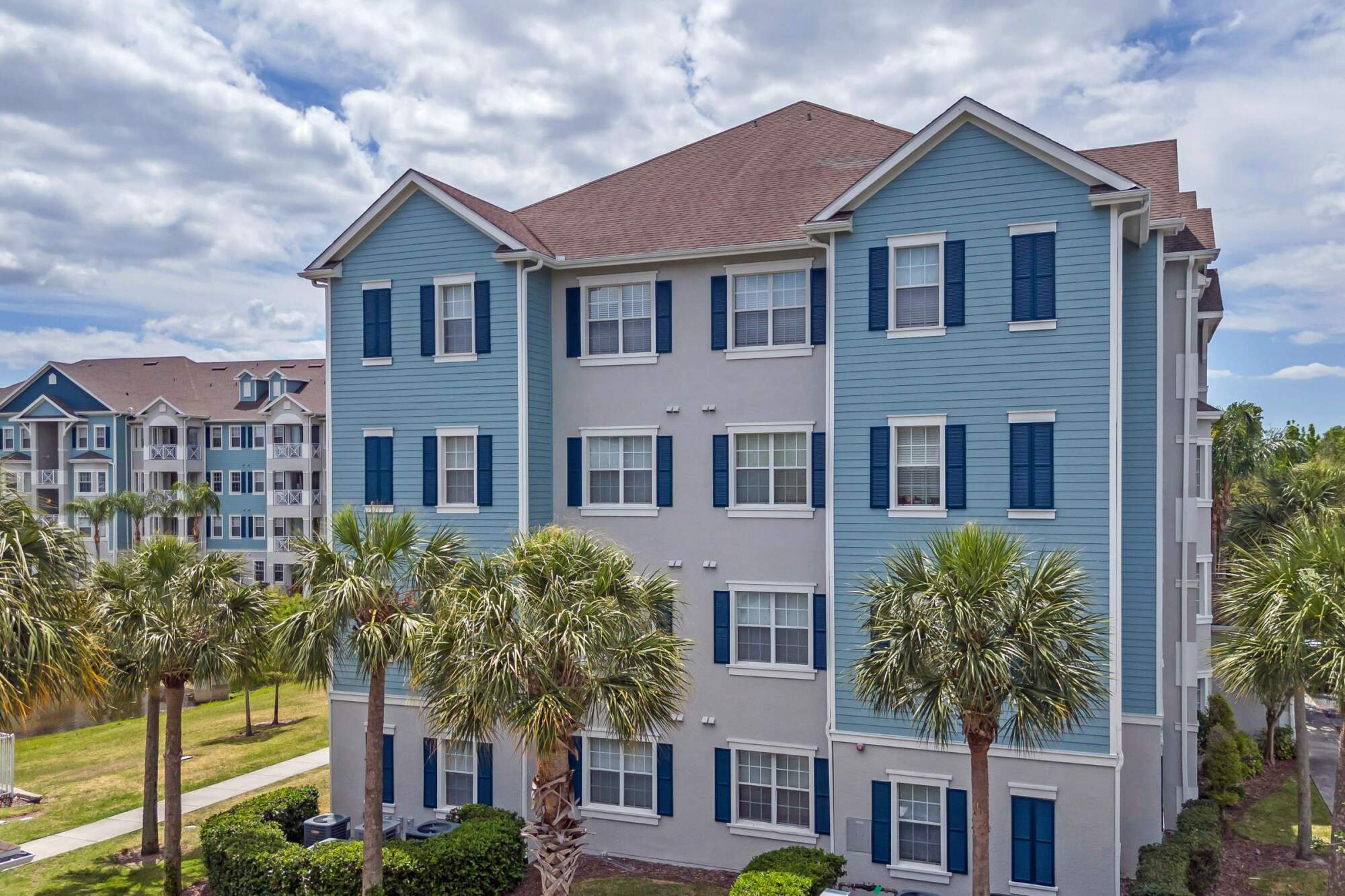 Four-story blue and white apartment building with palm trees in front under a partly cloudy sky.