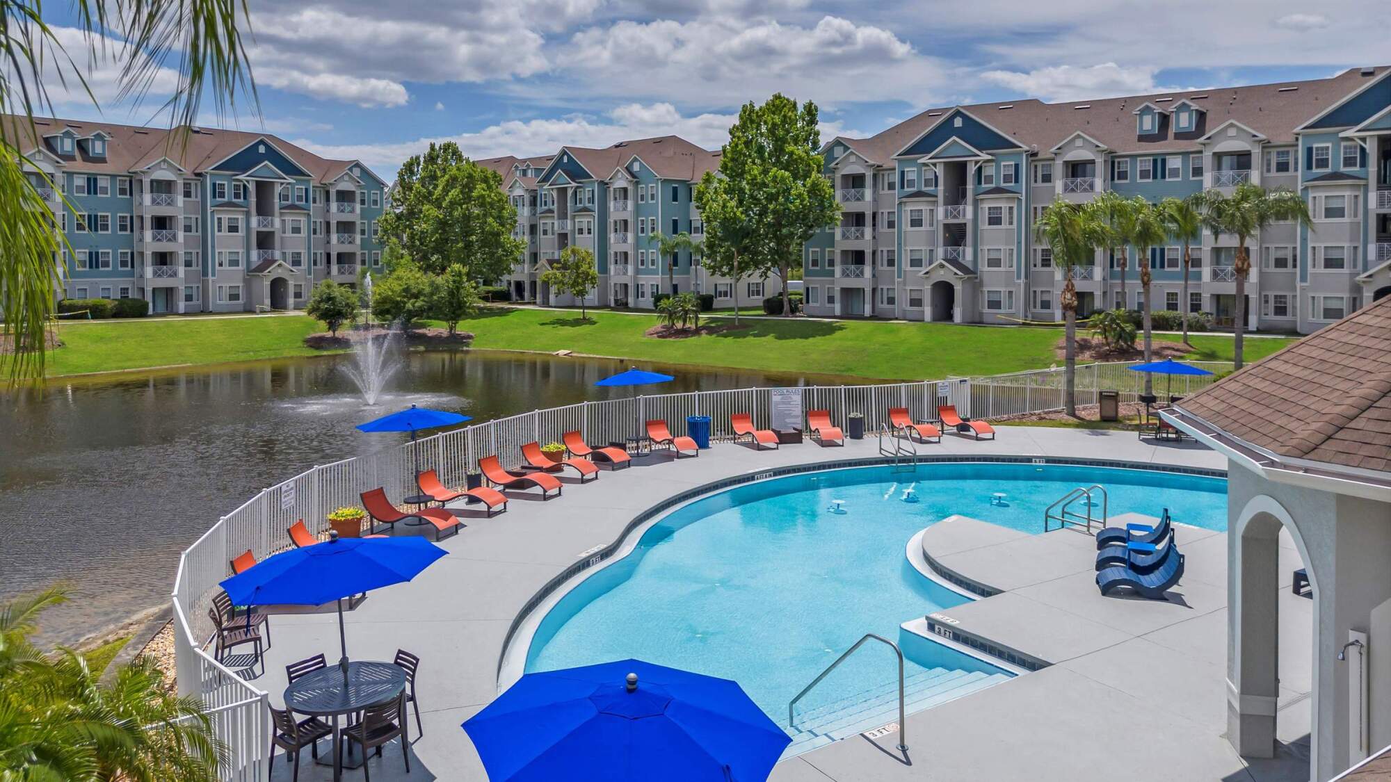 Curved outdoor pool with red lounge chairs, blue umbrellas, and pond with fountain near apartment buildings.