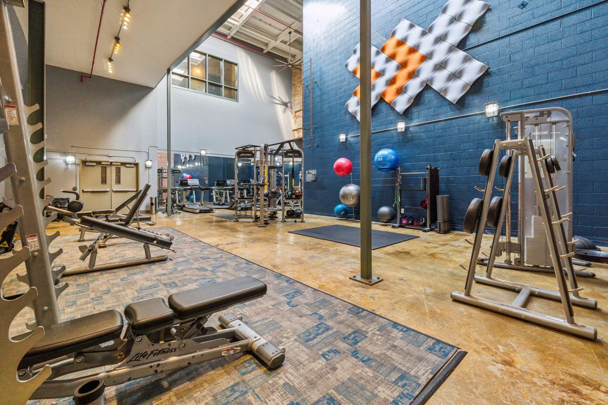 Modern gym with weight benches, exercise machines, stability balls, and wall art under high ceilings and bright lighting.