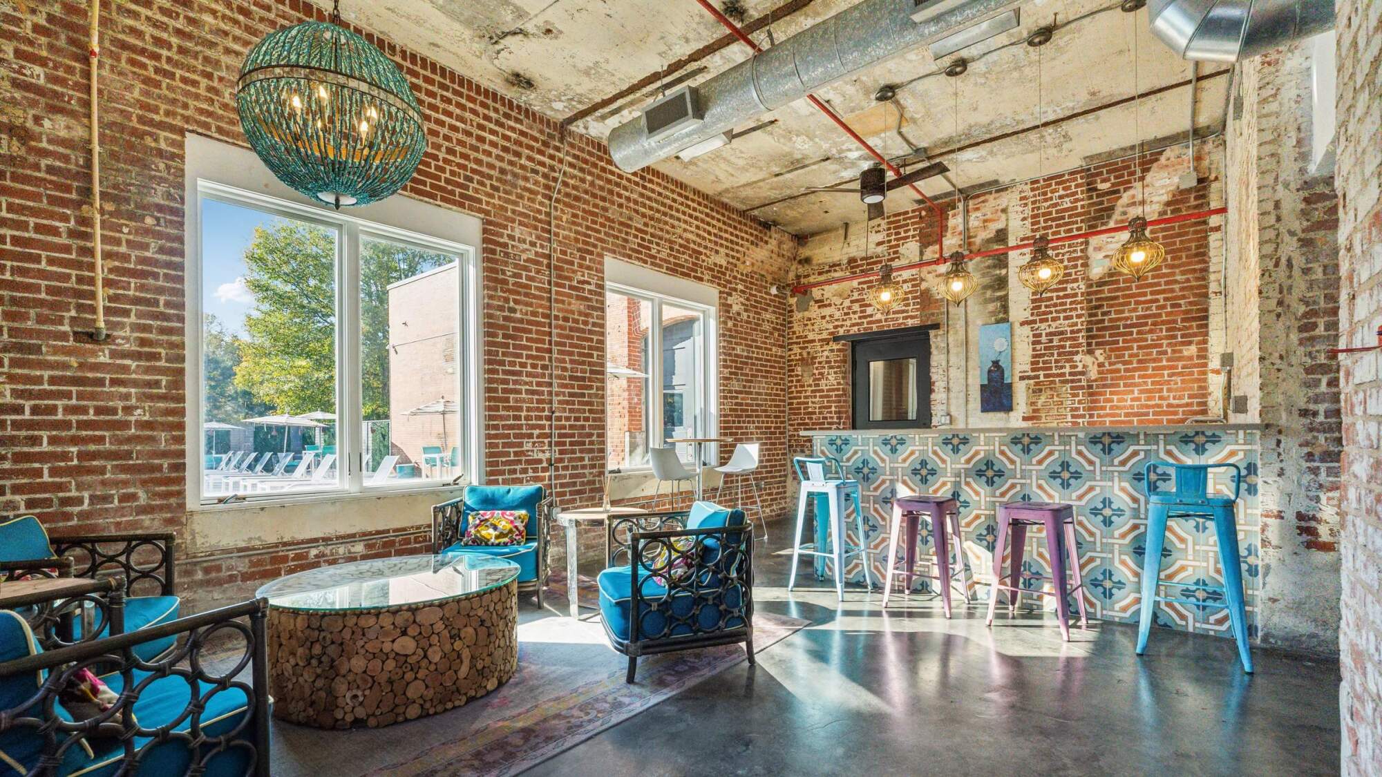 Modern lounge with exposed brick walls, colorful chairs, patterned bar, and large windows letting in sunlight.