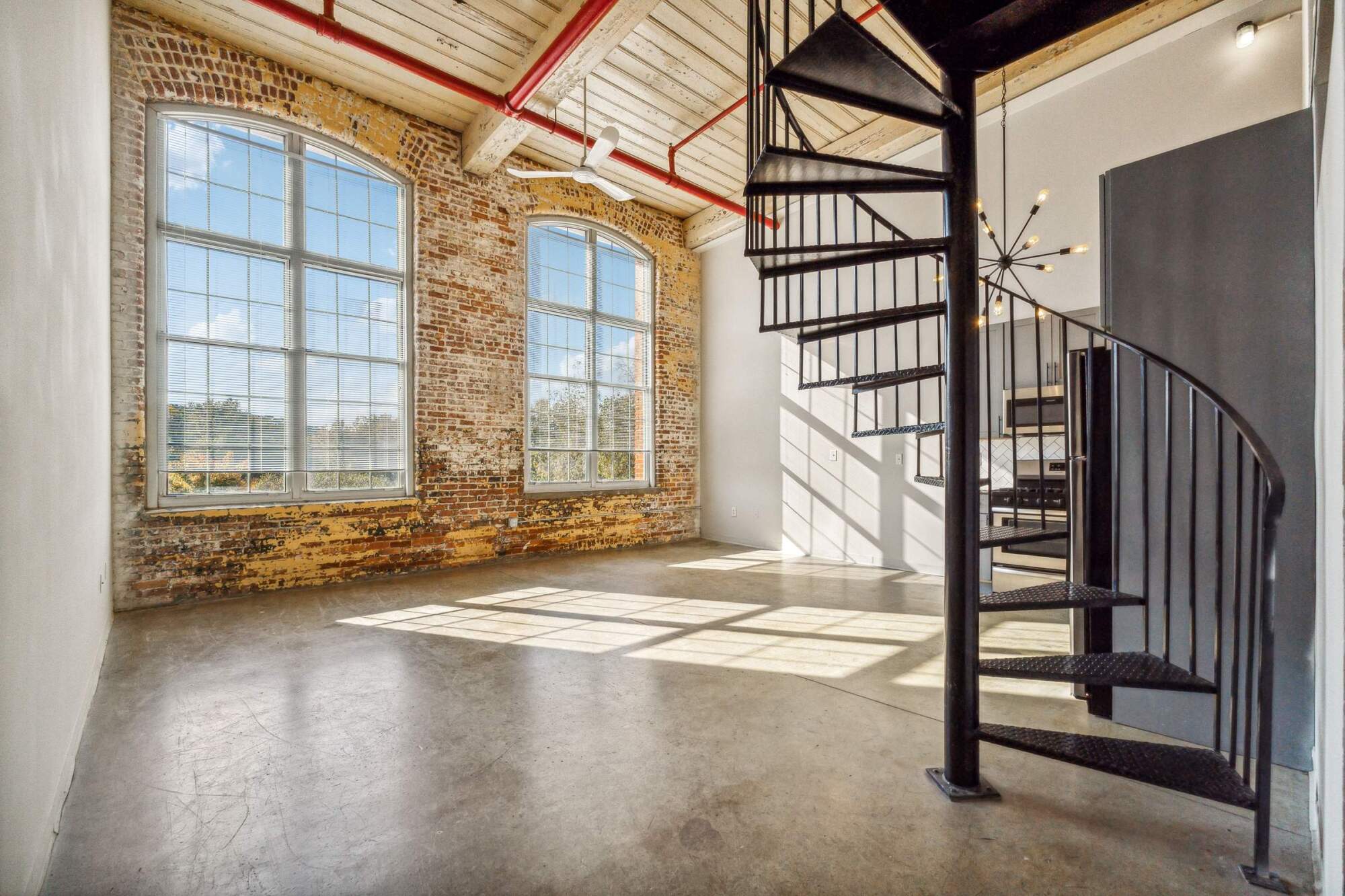 Sunlit loft with large arched windows, exposed brick walls, and a black spiral staircase.