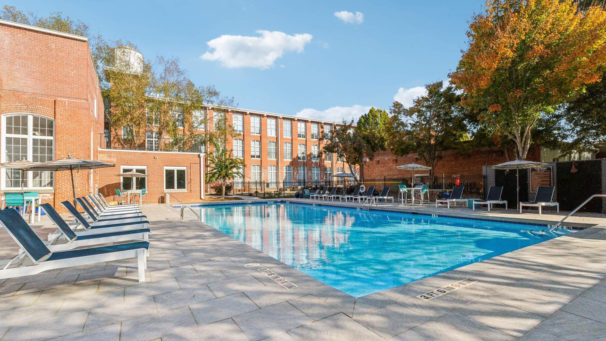 Outdoor swimming pool with lounge chairs, beside a brick building under a blue sky with scattered clouds.