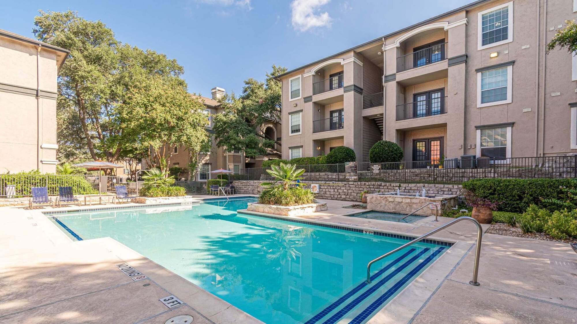 Outdoor swimming pool with clear water, surrounded by apartment buildings and lush greenery on a sunny day.