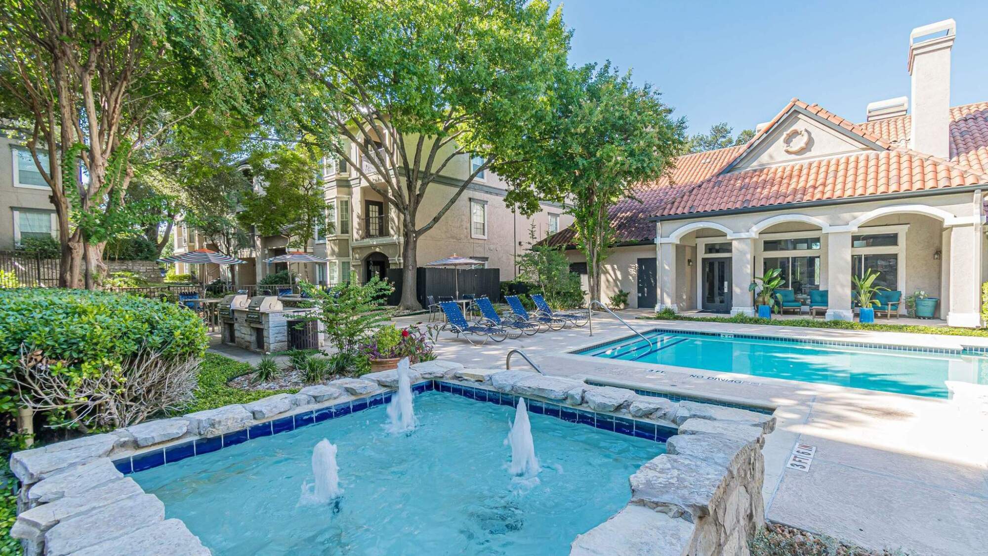 Outdoor pool area with lounge chairs, hot tub with fountains, and surrounding trees near apartment buildings.