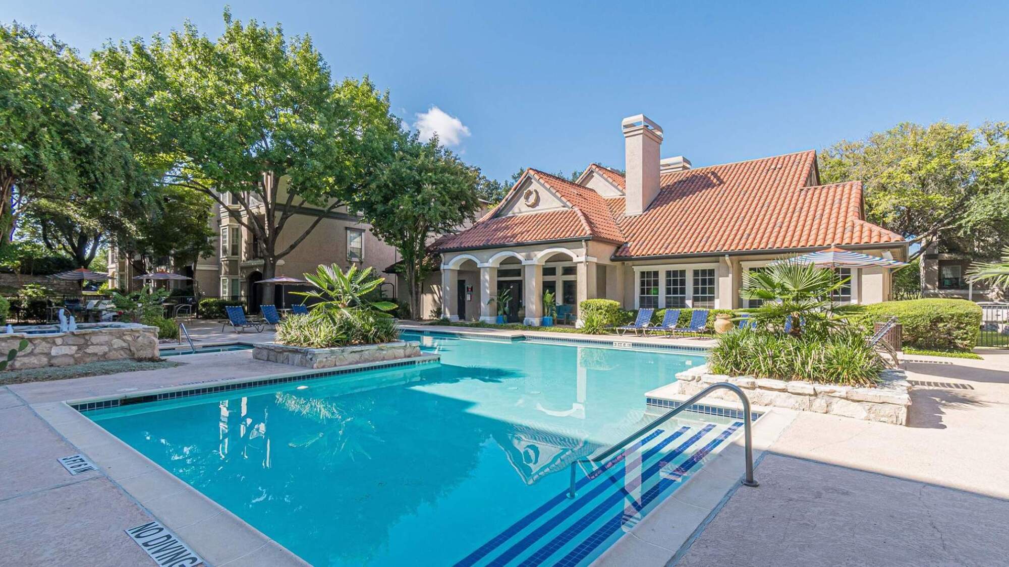 Bright outdoor pool with lounge chairs, greenery, and a building with a red-tiled roof in the background.
