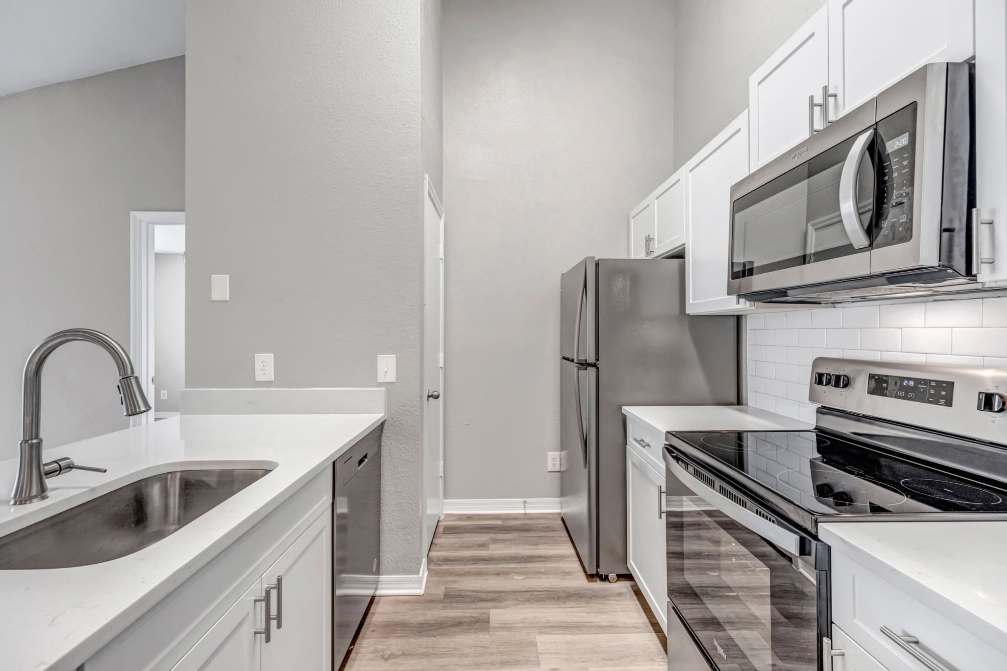 Galley-style apartment kitchen with white cabinets, quartz countertops, stainless steel appliances, white subway tile backsplash, deep stainless sink, and light wood-look flooring.
