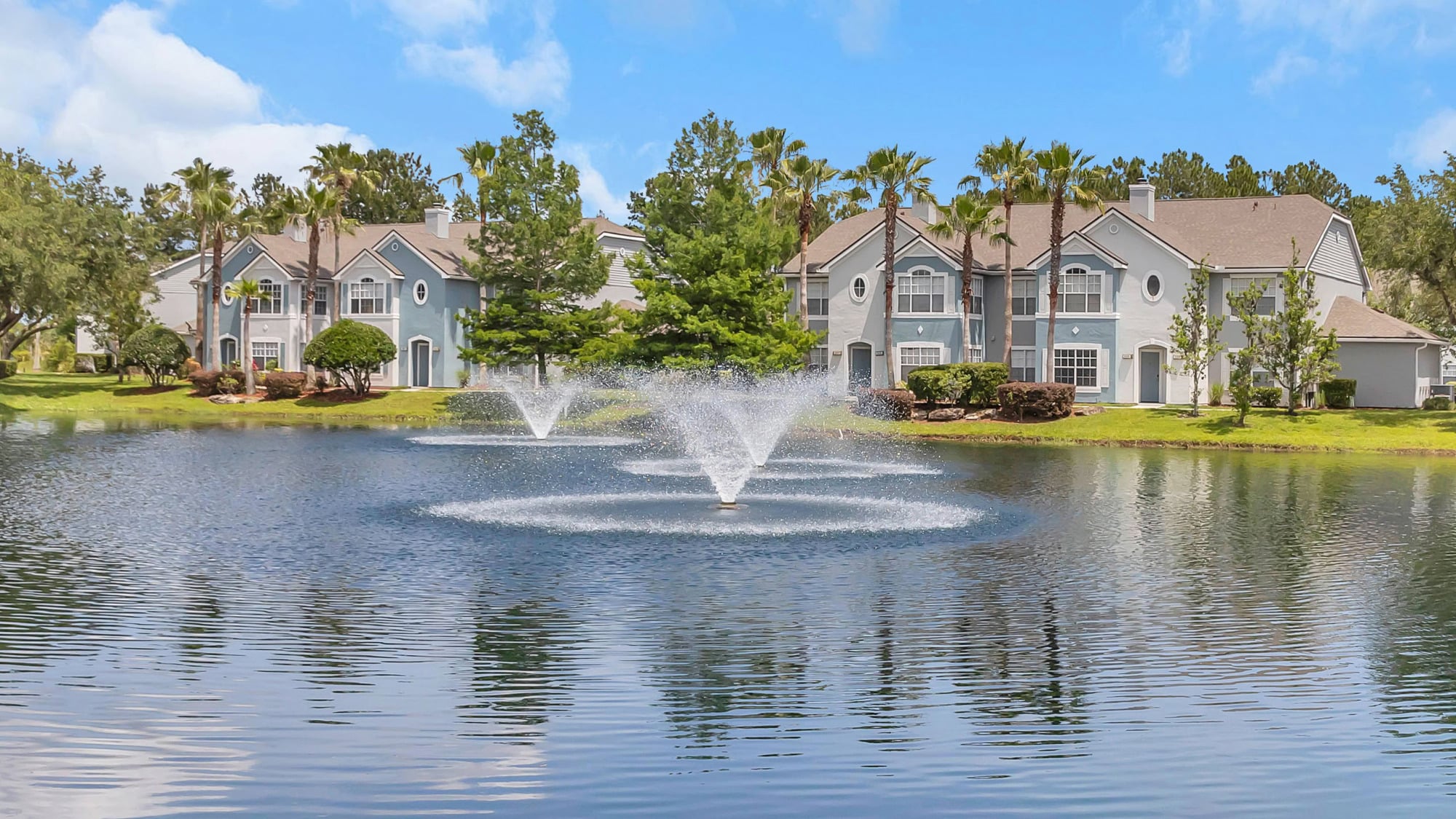 Picturesque community lake with a tall water fountain, mirror-like reflections, and stately townhome buildings framed by palm trees and lush landscaping in the background.