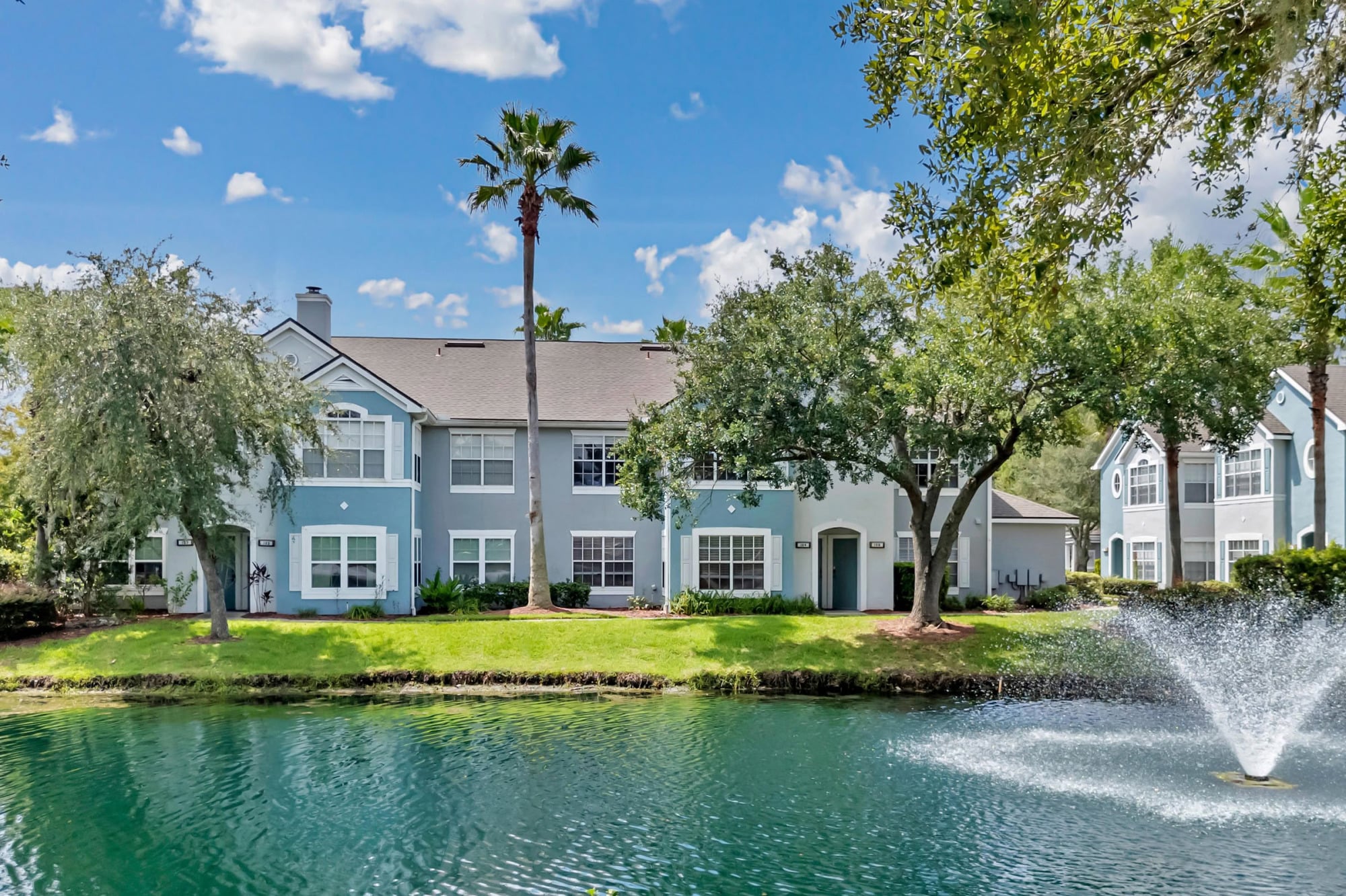 Lakefront view of a coastal blue and white townhome community with peaked rooflines, towering palm trees, a manicured lawn, and a fountain rippling across calm water.