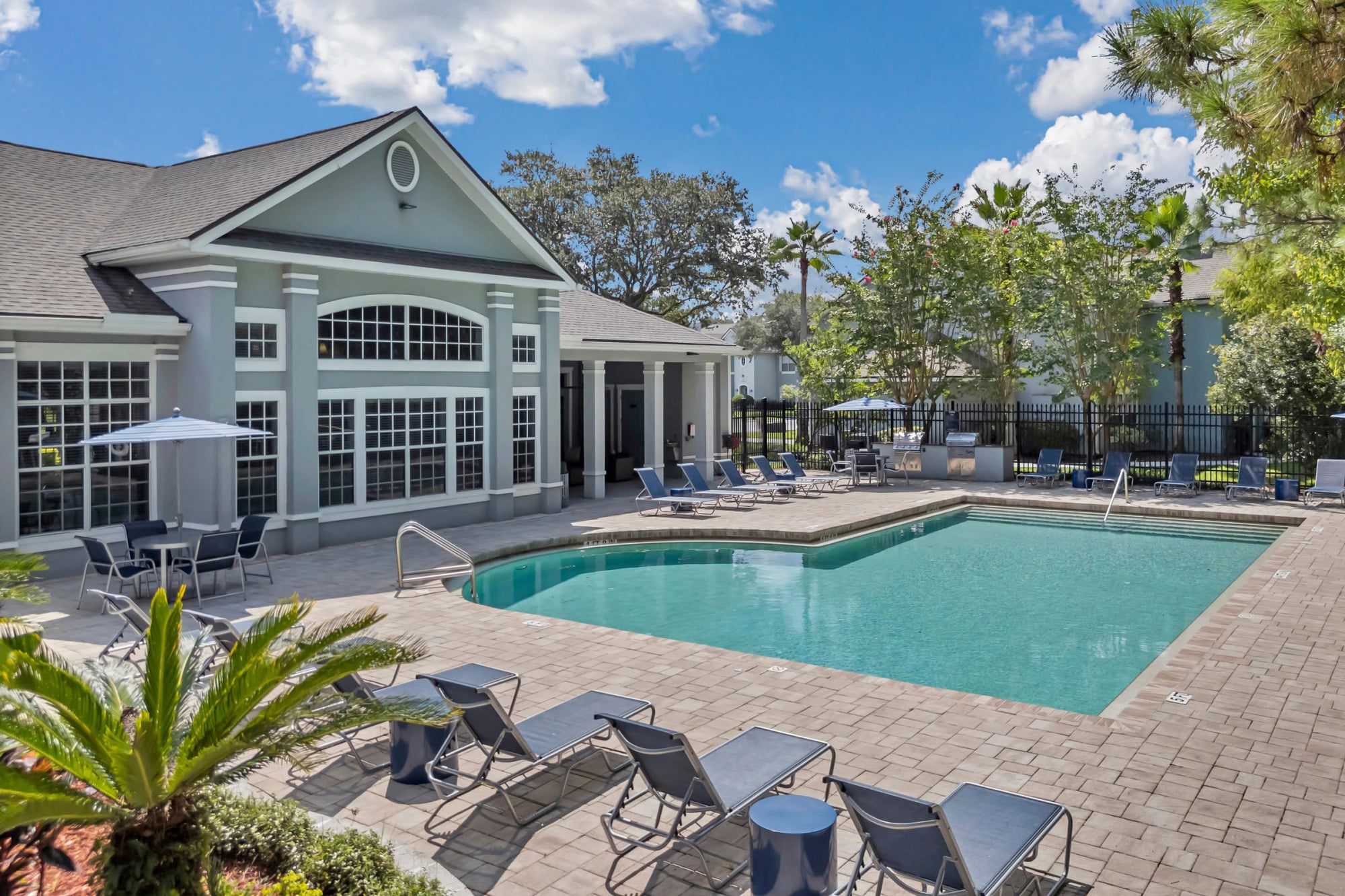 Tropical resort-style pool with paver patio, lounge chairs, an umbrella-shaded dining set, palm trees, and the gabled clubhouse exterior under a partly cloudy sky.
