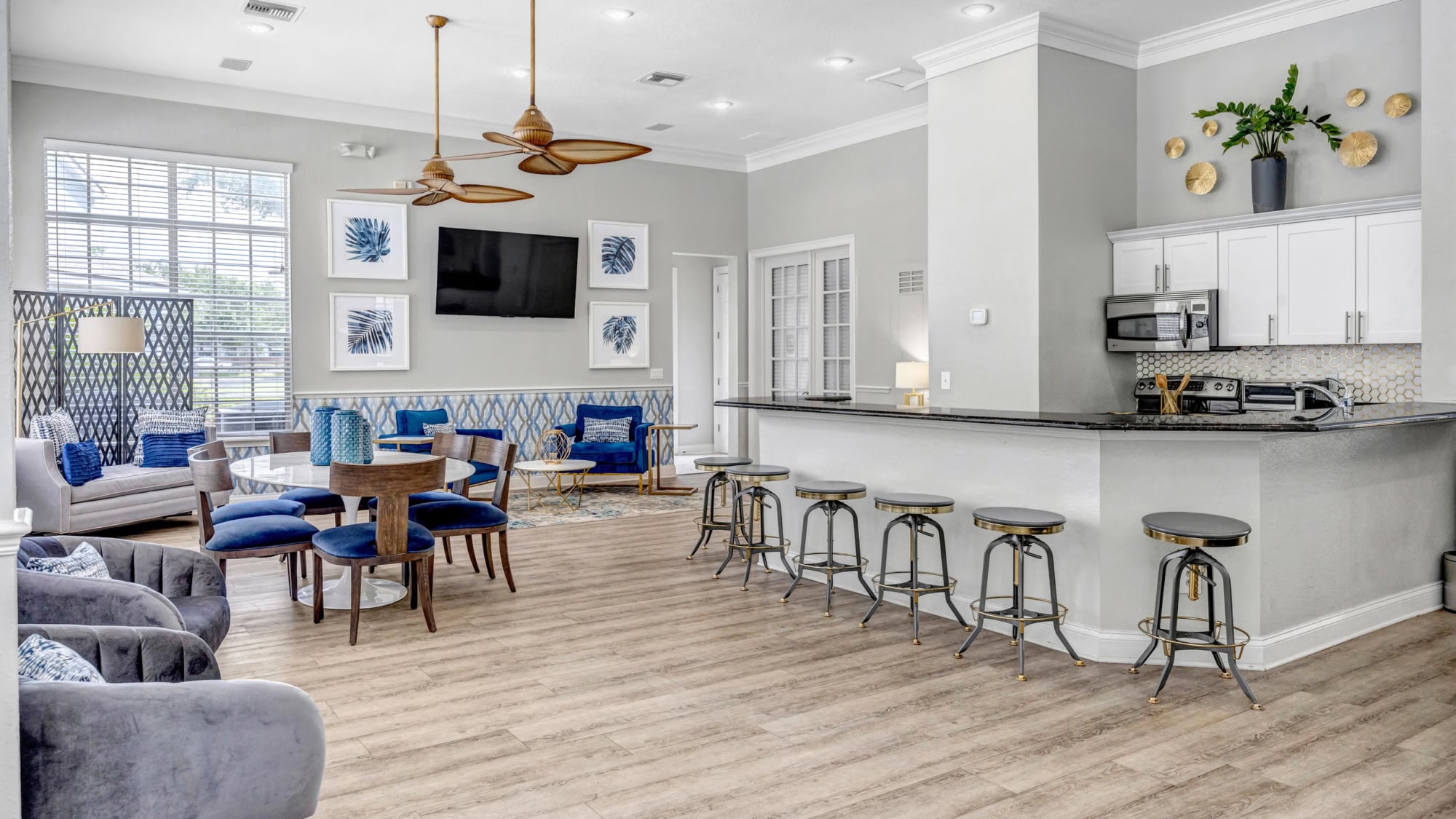 Coastal-inspired clubhouse lounge with light wood floors, blue and gray seating, a wall-mounted TV, decorative palm-print art, ceiling fans, and a kitchenette with industrial barstools.