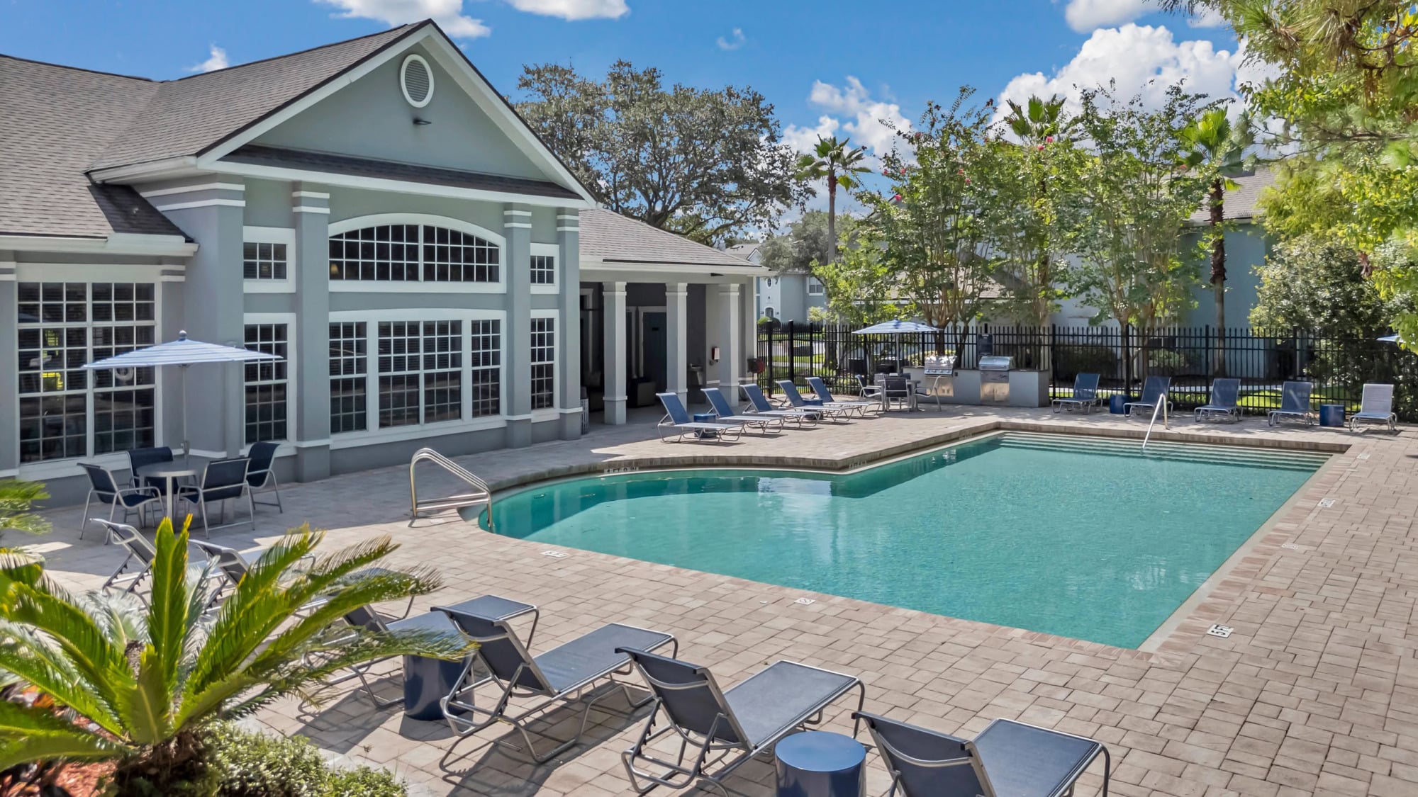 Resort-style swimming pool surrounded by paver decking and lounge chairs, with the elegant gabled clubhouse, palm trees, and a breezy umbrella seating area in the background.