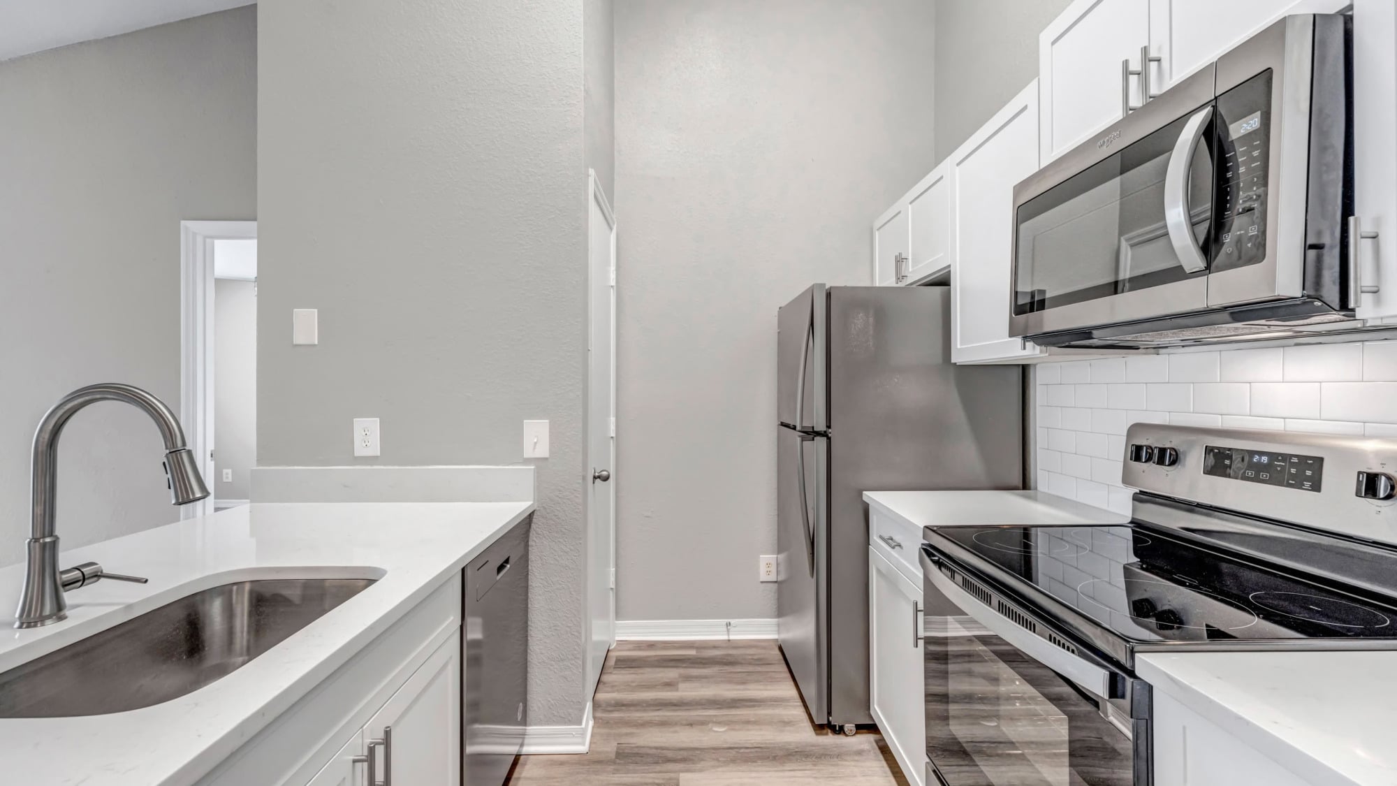 Bright apartment kitchen with white shaker cabinets, white quartz countertops, stainless steel appliances, white subway tile backsplash, and warm wood-look flooring beneath soaring ceilings.