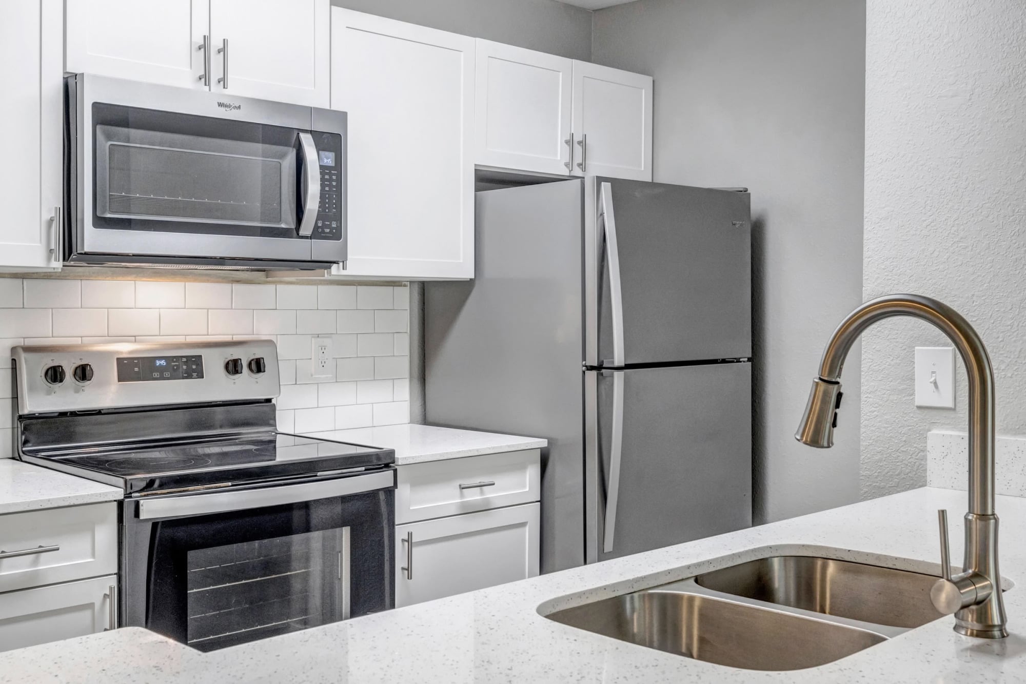 Close-up of an apartment kitchen with white shaker cabinetry, stainless steel microwave, range, and refrigerator, white subway tile backsplash, quartz counters, and a brushed-nickel pull-down faucet over a stainless sink.