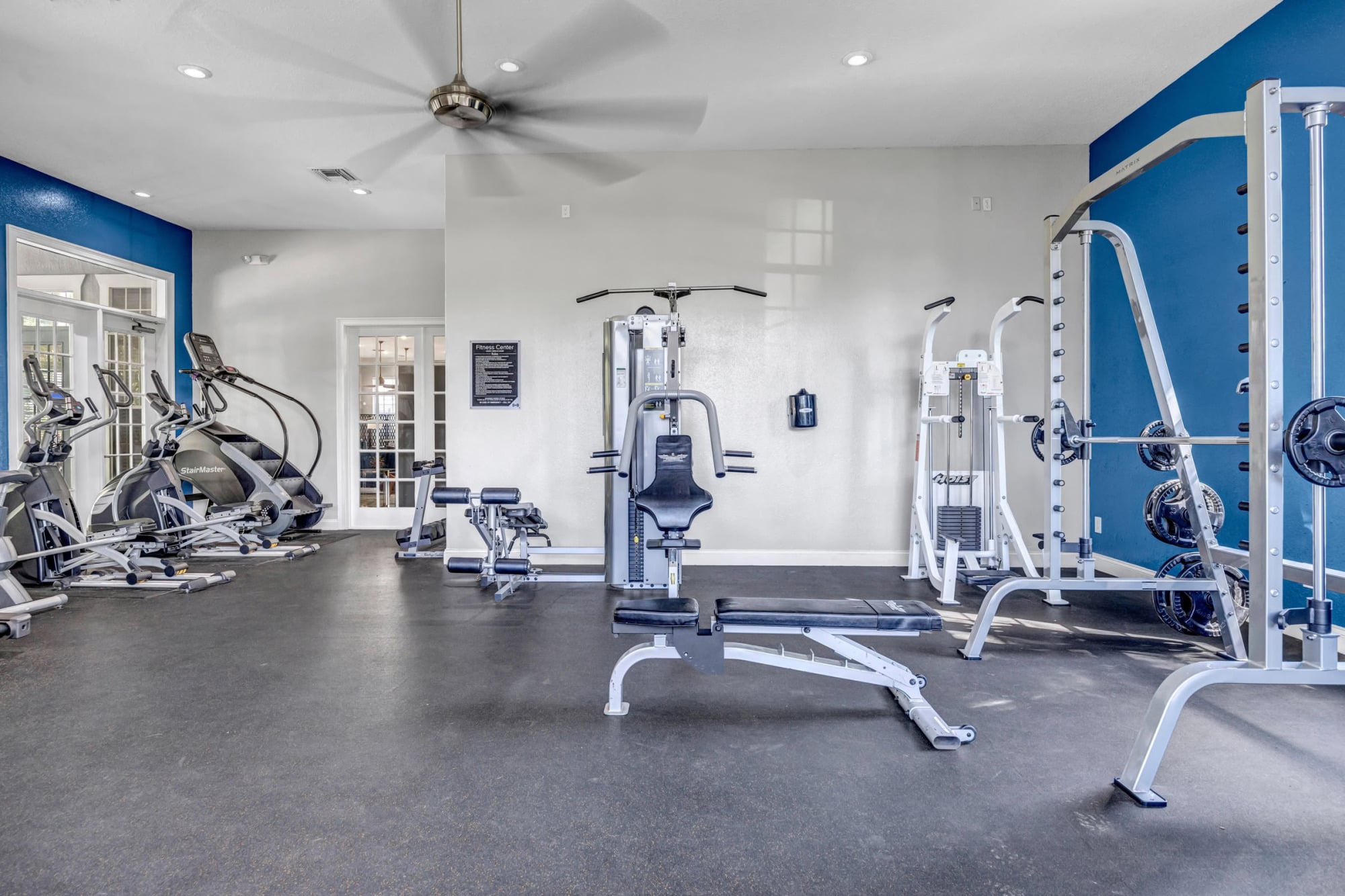 Open fitness studio with cardio equipment along one wall, weight machines and a bench press, ceiling fan, and a blue accent wall providing a coastal vibe.