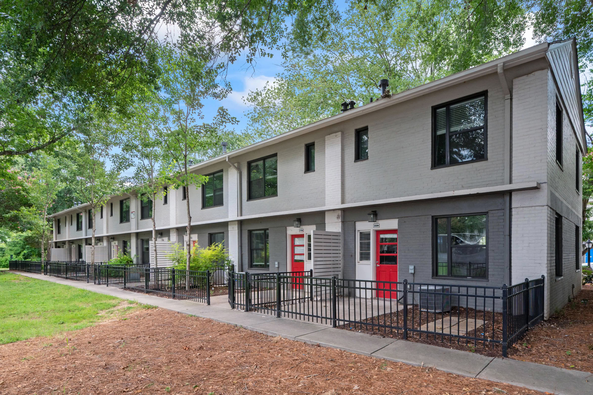 Updated two-story gray apartment building with red front doors, black wrought-iron fenced patios, mature trees, and a wood-mulched walkway leading along the building.