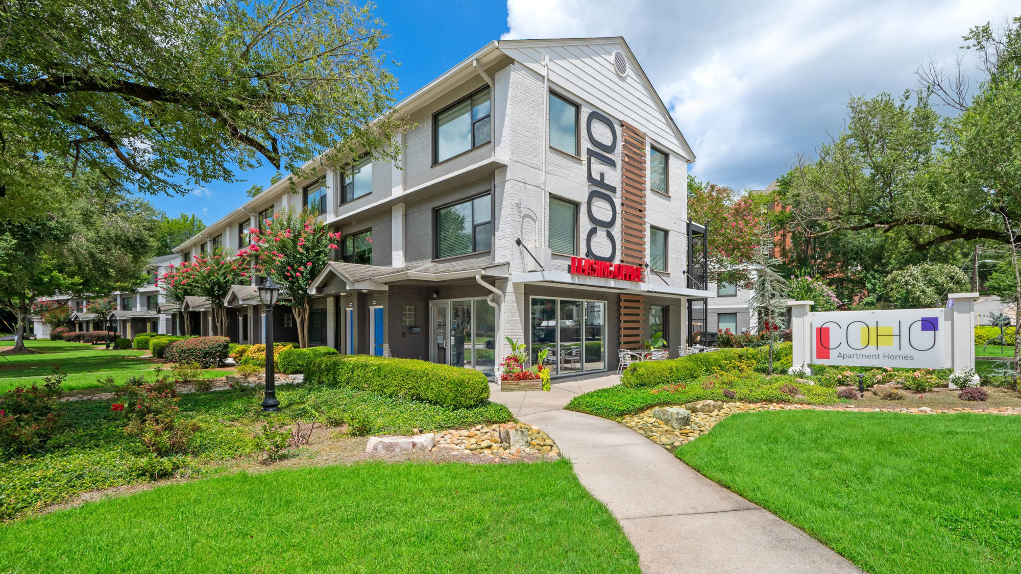 Three-story modern apartment building with white and gray exterior, large CoHo branding, glass entry doors, lush landscaped lawn, and the CoHo Apartment Homes signage near the walkway.