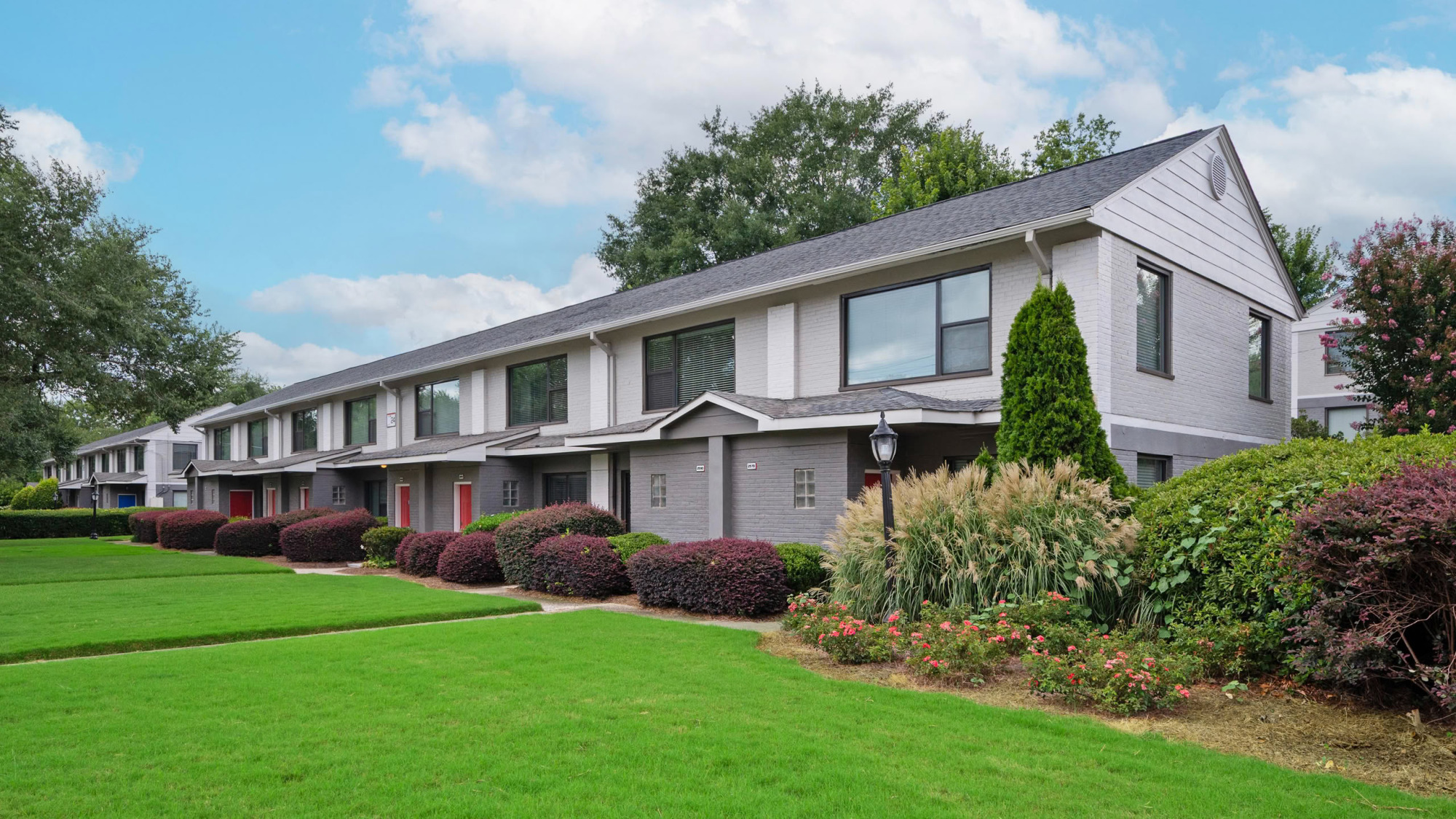 Long row of two-story gray brick townhomes with peaked rooflines, red and dark front doors, manicured shrubs, and a neat green lawn under a partly cloudy sky.