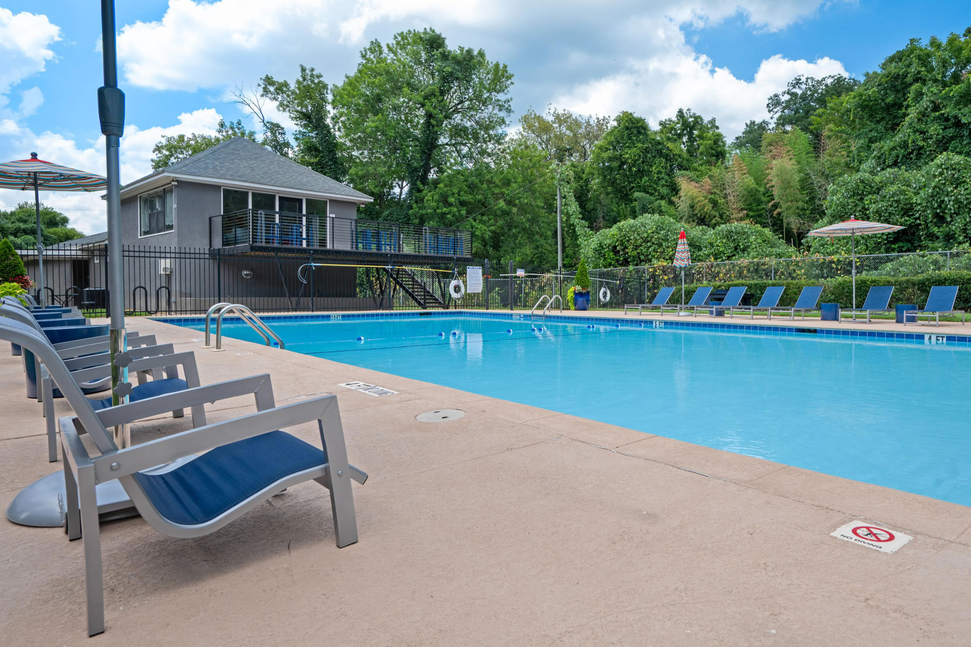 Inviting community pool with crystal blue water, lounge chairs lined along paver decking, vibrant umbrellas, and a contemporary gray pool house surrounded by trees.