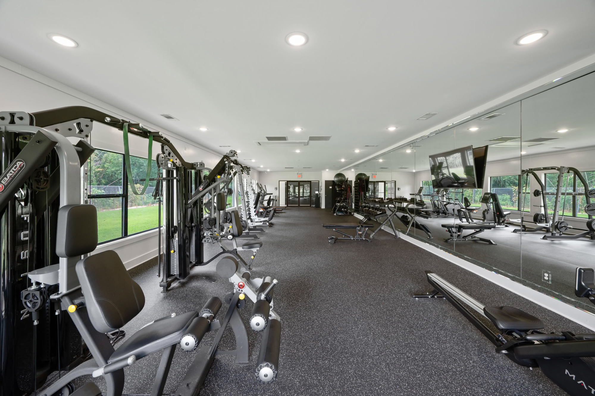 Long fitness studio featuring a multi-station cable machine, weight benches, mirrored accent wall, polished black floors, and tall windows with garden views.