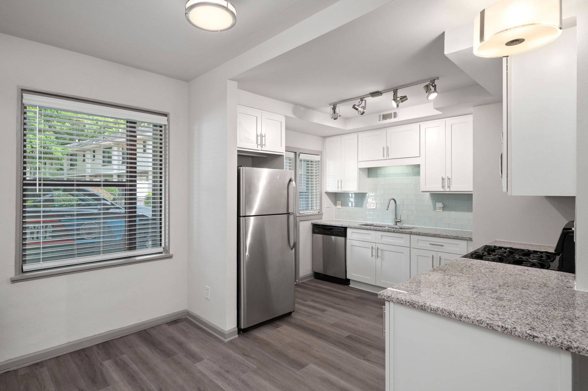 Open kitchen and dining area featuring white shaker cabinets, light blue glass subway tile backsplash, granite countertops, stainless steel appliances, modern lighting, and a bright window with neighborhood views.