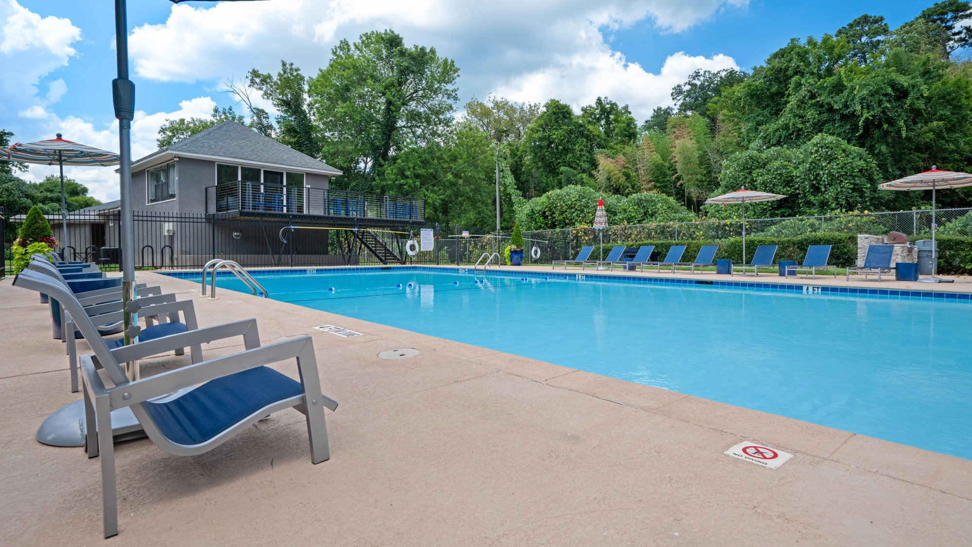 Outdoor swimming pool surrounded by lounge chairs and umbrellas, with a charcoal-gray pool house and lush green trees framing a partly cloudy sky.