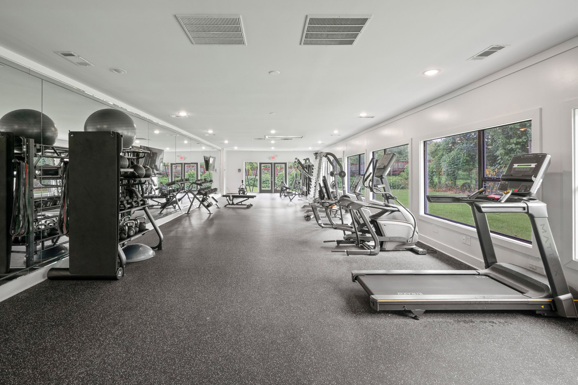 Bright two-zone gym with treadmills and ellipticals lining the windowed wall, free weight rack with stability balls, and a mirrored wall reflecting the equipment.