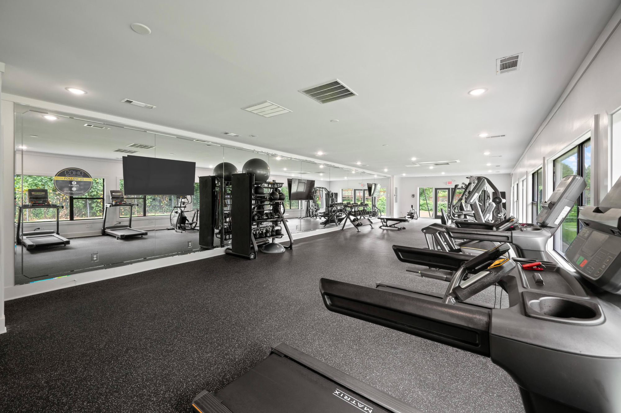 Modern fitness center with row of treadmills facing wall-mounted TVs, mirrored wall, dumbbell rack, weight bench, and large windows letting in natural light.