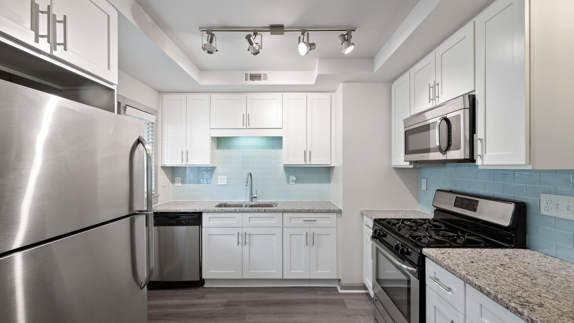 Updated apartment kitchen with bright white shaker cabinets, light blue glass tile backsplash, granite countertops, stainless steel appliances, gas range, and modern track lighting overhead.