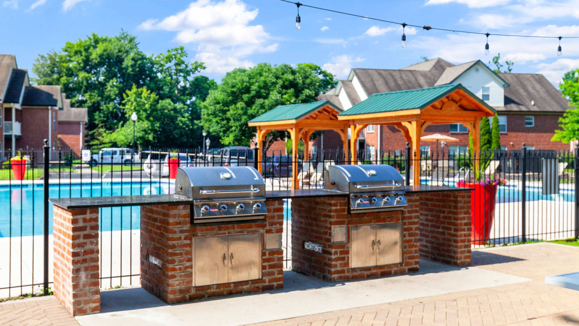 Outdoor brick grilling station with two stainless steel barbecue grills, string lights overhead, and a wooden pergola and pool visible in the background of the community courtyard.