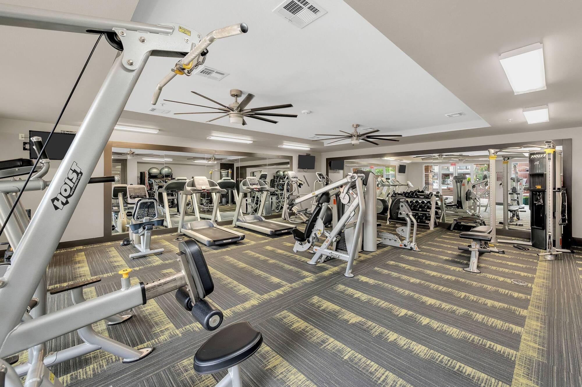 Modern gym with cardio and strength equipment, large mirrors, and ceiling fans on a patterned floor.