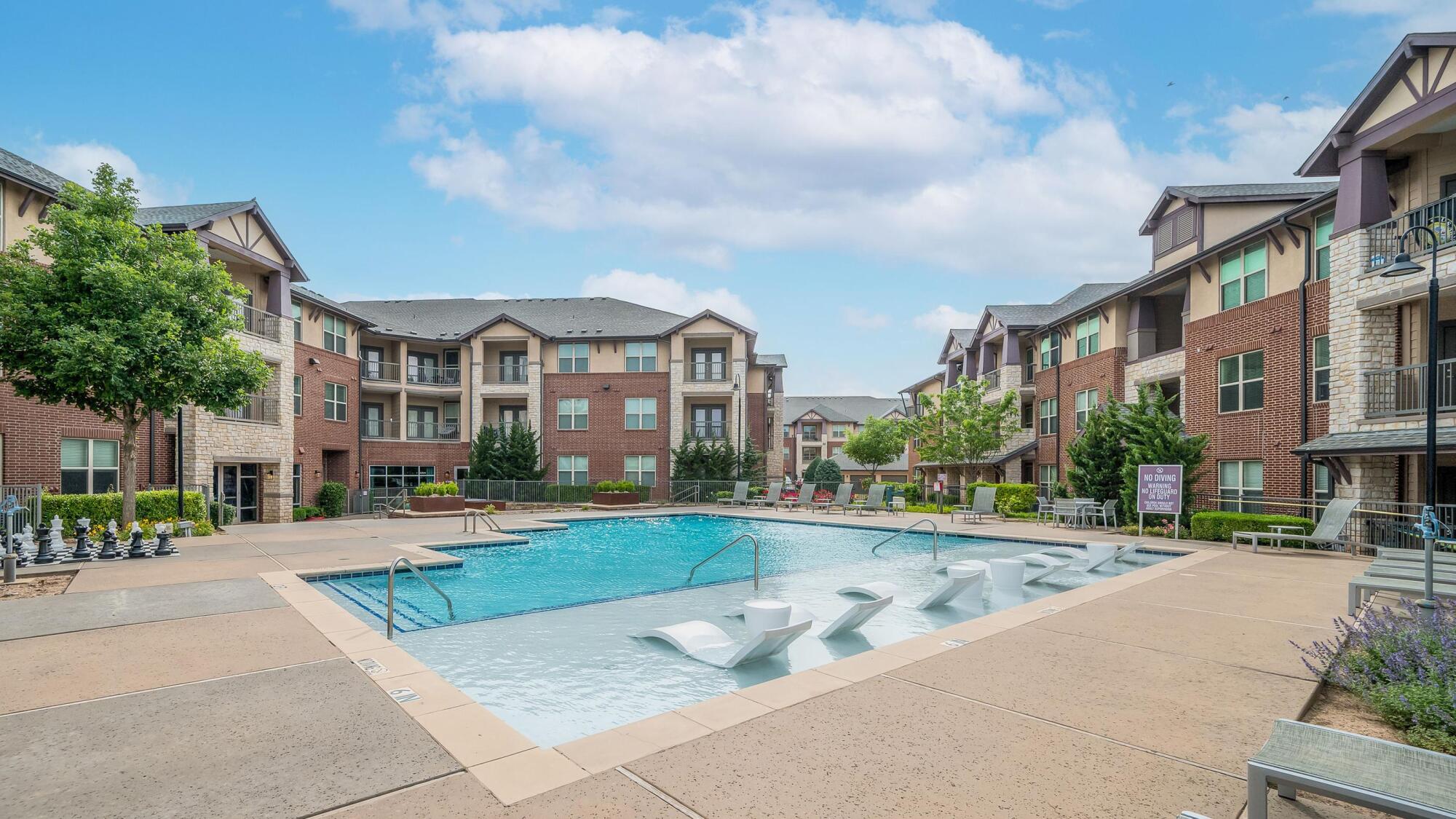 Outdoor apartment pool area with lounge chairs and trees, surrounded by multi-story brick buildings on a sunny day.