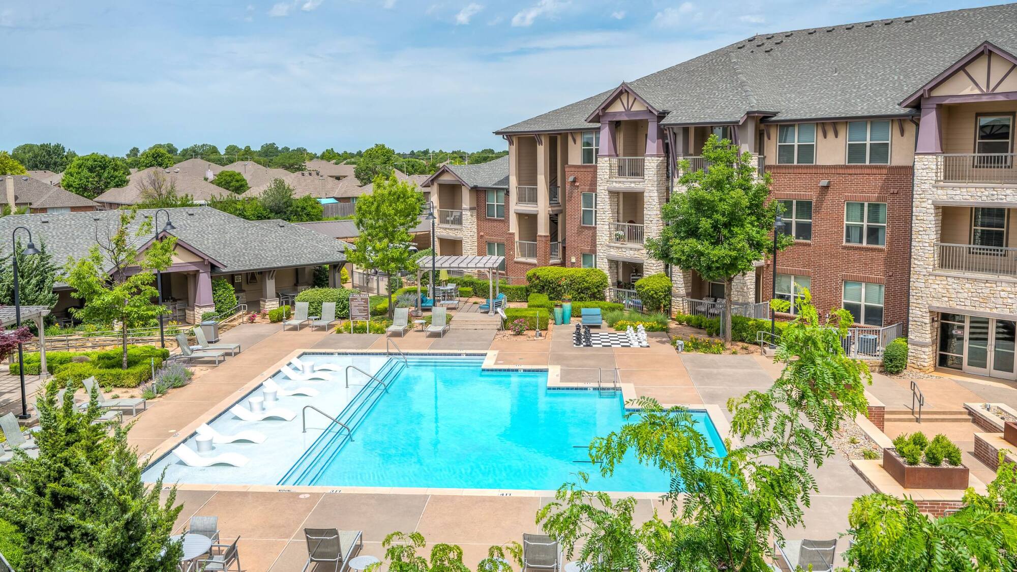 Apartment complex with a large outdoor pool, lounge chairs, and landscaped greenery on a sunny day.