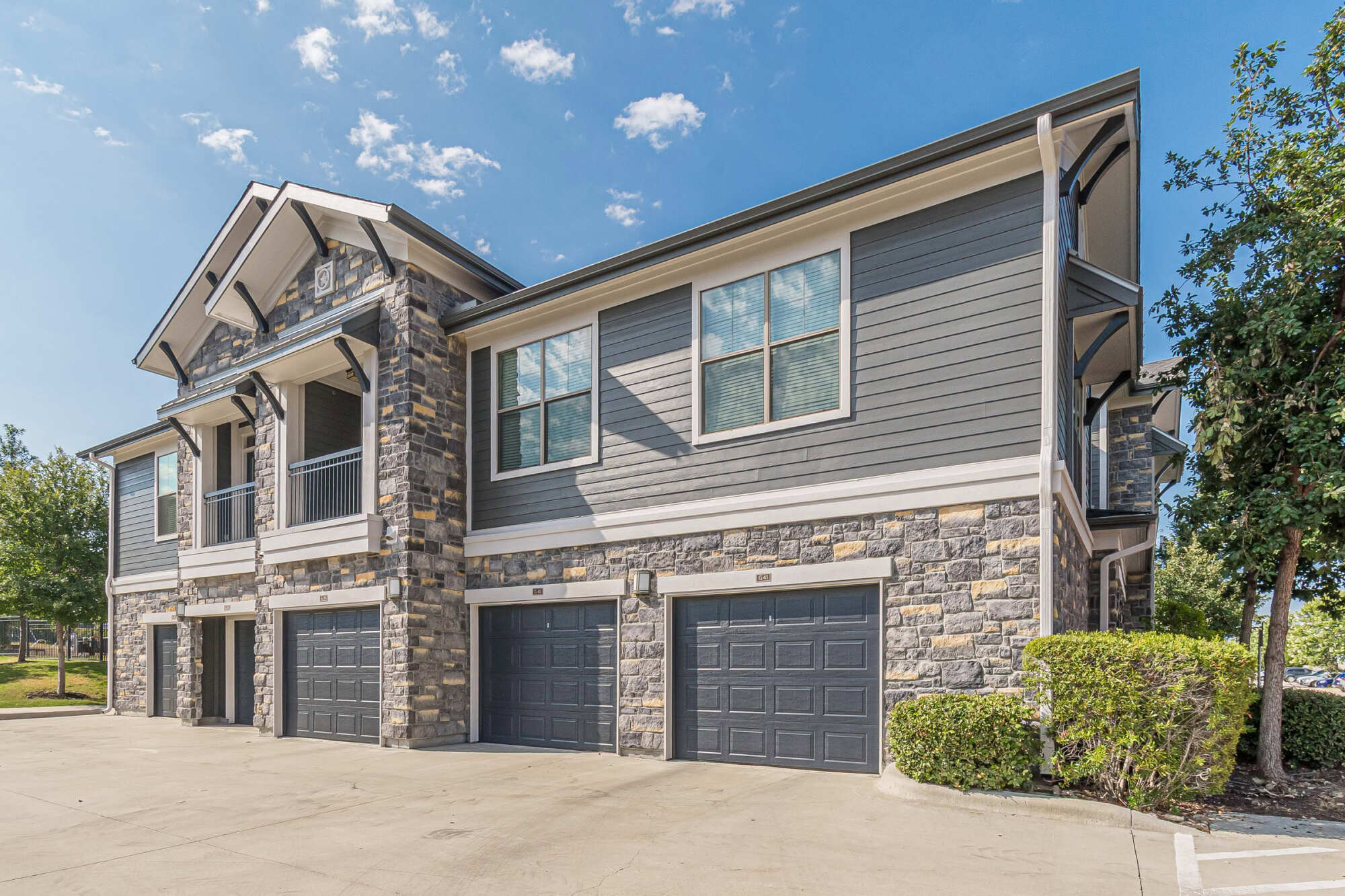 Two-story apartment building featuring stone veneer accents, gray horizontal siding, attached private garages, peaked gables, and manicured landscaping under a clear blue sky.