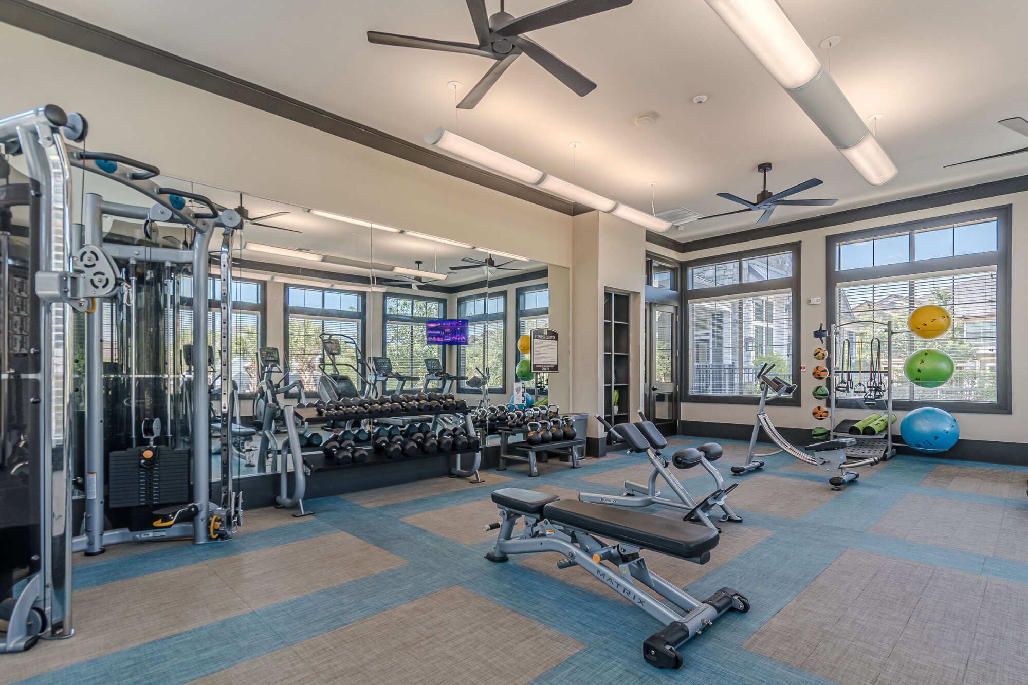 Bright fitness center with a cable weight machine, full dumbbell rack, adjustable workout bench, mirrored wall, large windows, and overhead ceiling fans illuminating the workout space.