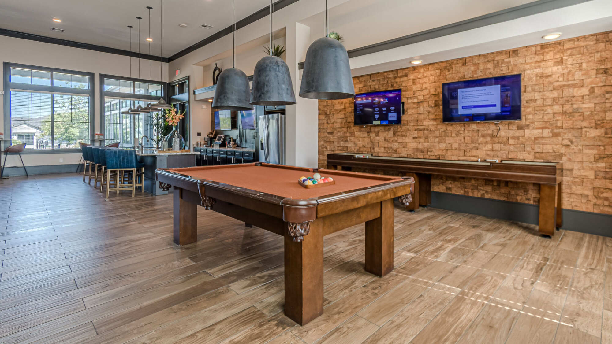Resident clubroom featuring a billiards table, shuffleboard table, brick accent wall with mounted TVs, industrial pendant lighting, and a bar area near floor-to-ceiling windows.