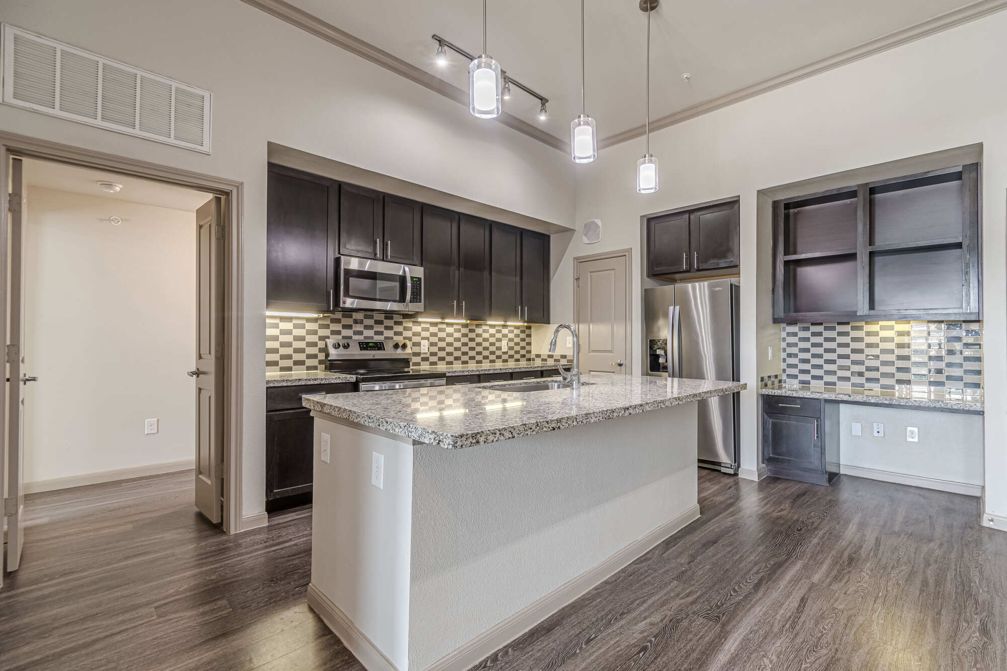 Spacious apartment kitchen with high ceilings, dark espresso cabinets, granite-topped island, stainless appliances, mosaic glass backsplash, modern pendant lights, and a built-in desk nook.