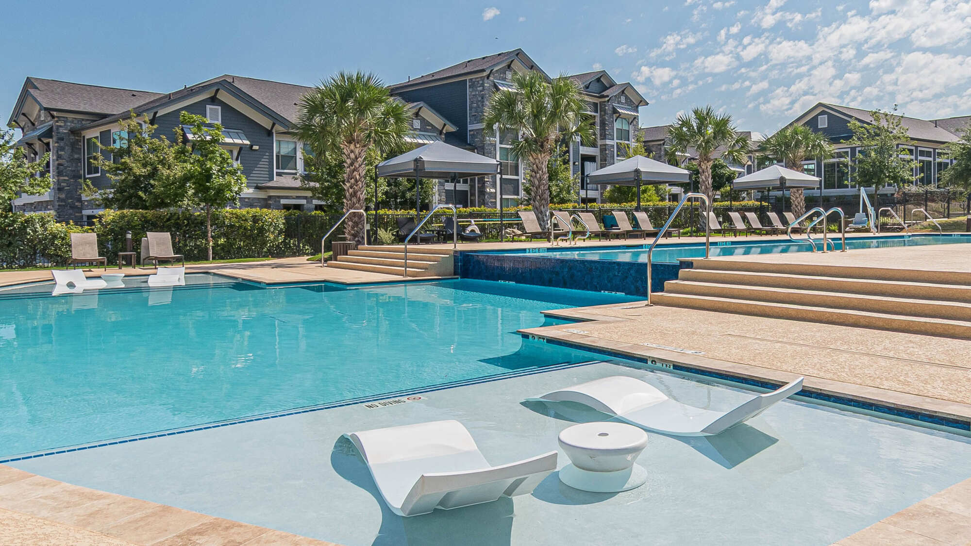 Resort-style swimming pool with white in-water tanning lounges, palm trees, gray umbrellas, and a multi-story apartment community with peaked gabled roofs in the background.