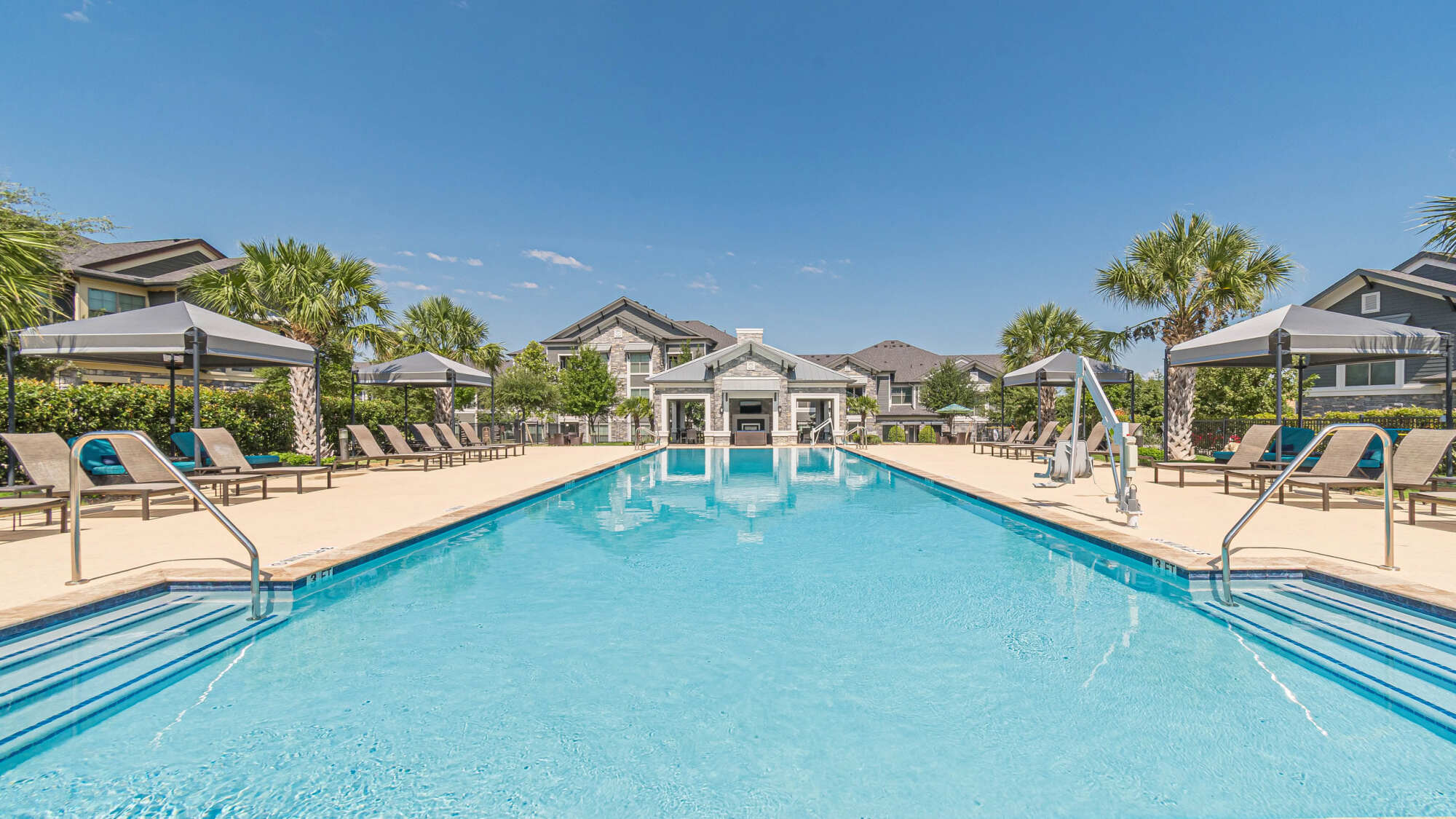 Large rectangular community pool flanked by shaded cabanas, palm trees, lounge chairs, and a traditional-style clubhouse with peaked rooflines and a Texas Hill Country feel.