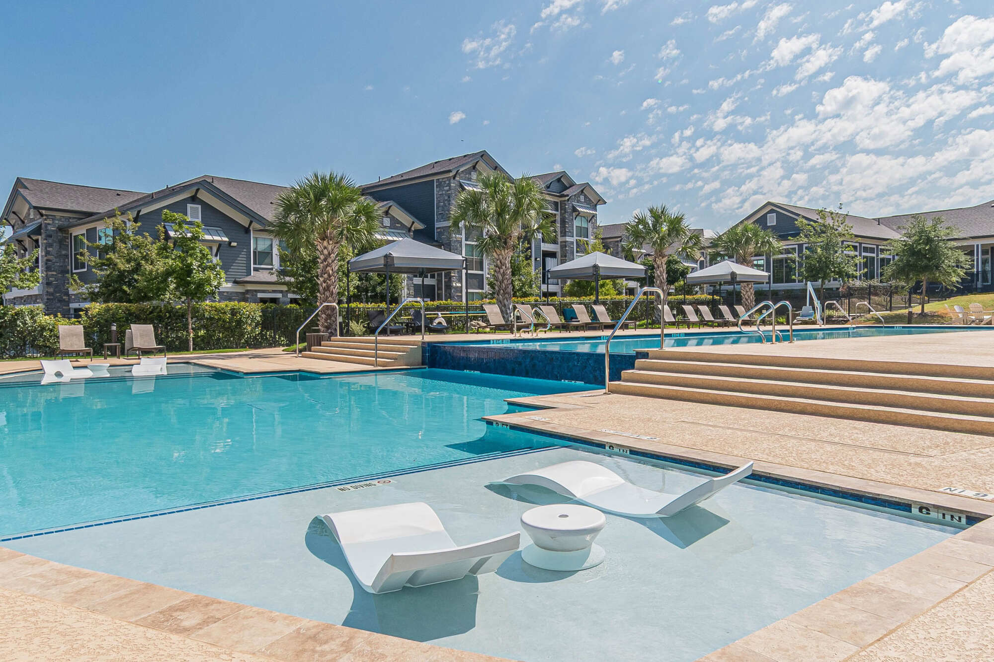 Sparkling resort-style pool with floating white tanning lounges, palm trees, paver decking, gray umbrellas with lounge chairs, and modern apartment buildings under a bright blue sky.