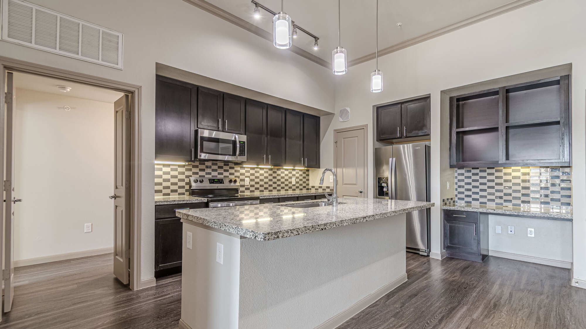 Open-concept apartment kitchen featuring dark espresso cabinetry, granite-topped center island, multi-tone tile backsplash, stainless steel appliances, modern pendant lighting, and a built-in office nook.