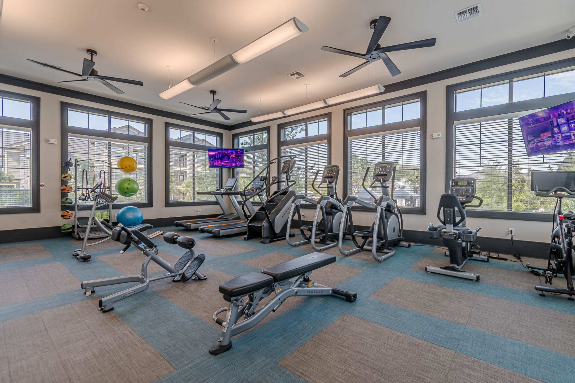 Modern cardio studio with treadmills, ellipticals, recumbent bikes, and rowing machines arranged on patterned blue and gray flooring, with stability balls and TVs mounted to the wall.
