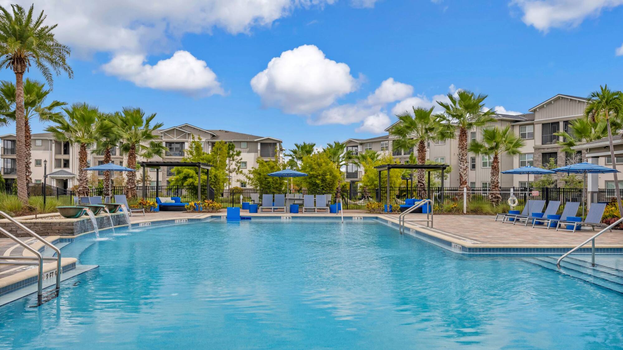 Large outdoor swimming pool with lounge chairs, umbrellas, palm trees, and apartment buildings in the background.