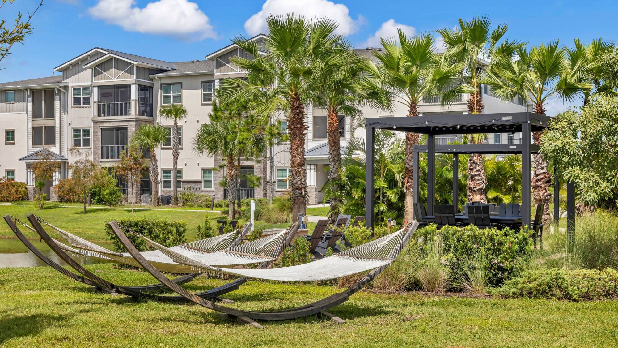Resort-style outdoor swimming pool with lounge chairs, umbrellas, and palm trees near modern apartments.